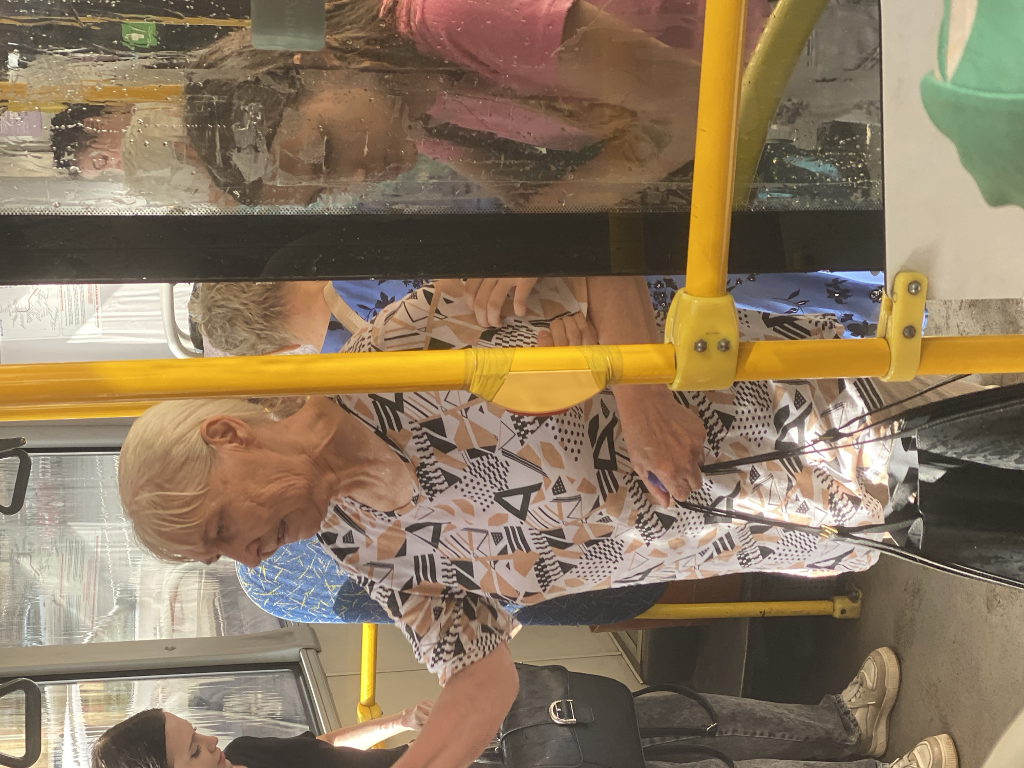The image depicts the interior of a public transportation vehicle, likely a bus, in Novokuznetsk, Russia. The scene is filled with passengers. In the foreground, an elderly woman with blonde hair, wearing a patterned blouse, is the primary subject. She is positioned centrally and appears to be interacting with someone or looking at something to her left. To her left, a younger woman with dark hair, wearing a black top and grey pants, is partially visible. She is carrying a black handbag with a buckle. In the background and to the right, other passengers are visible, some seen through the condensation-covered windows. The interior of the bus features yellow metal poles and handles, characteristic of public transport. The windows are wet, suggesting it might be raining or humid. The overall atmosphere is typical of everyday public transit in an urban environment.