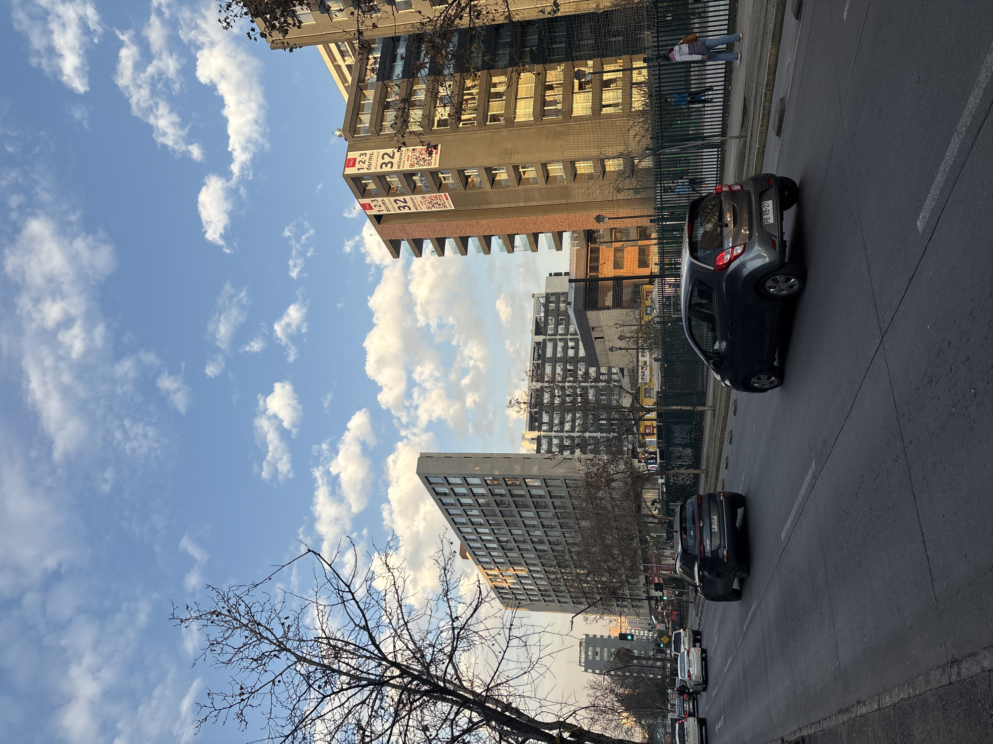 This is a street view in Santiago, Chile, on a partly cloudy day, likely in the afternoon given the lighting. The image captures a busy street with multiple lanes of traffic. Several cars are visible, including a dark gray Kia Sportage and a smaller dark gray hatchback. In the background, there are tall apartment buildings with many windows, some displaying signs with promotional material for apartments, including the numbers "1-2-3," "32," and "Millones." Bare-branched trees line the street in the foreground and between the sidewalk and the road. Pedestrians are visible on the sidewalk behind a fence. The sky is blue with scattered white clouds. The architecture suggests an urban environment typical of a large city.