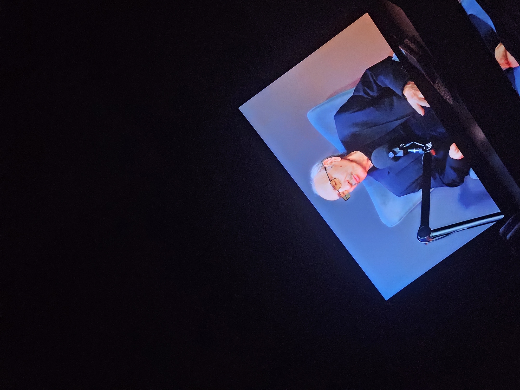 A man with a mustache and glasses is seated in a chair, wearing a suit jacket. A microphone is positioned in front of him. The scene is viewed on a screen, with a dark background. The screen is angled, displaying a portion of the man's upper body and the chair he is sitting in. The lighting suggests an indoor setting, likely a studio or a broadcast environment. The image is a digital photograph with a clear focus on the man.