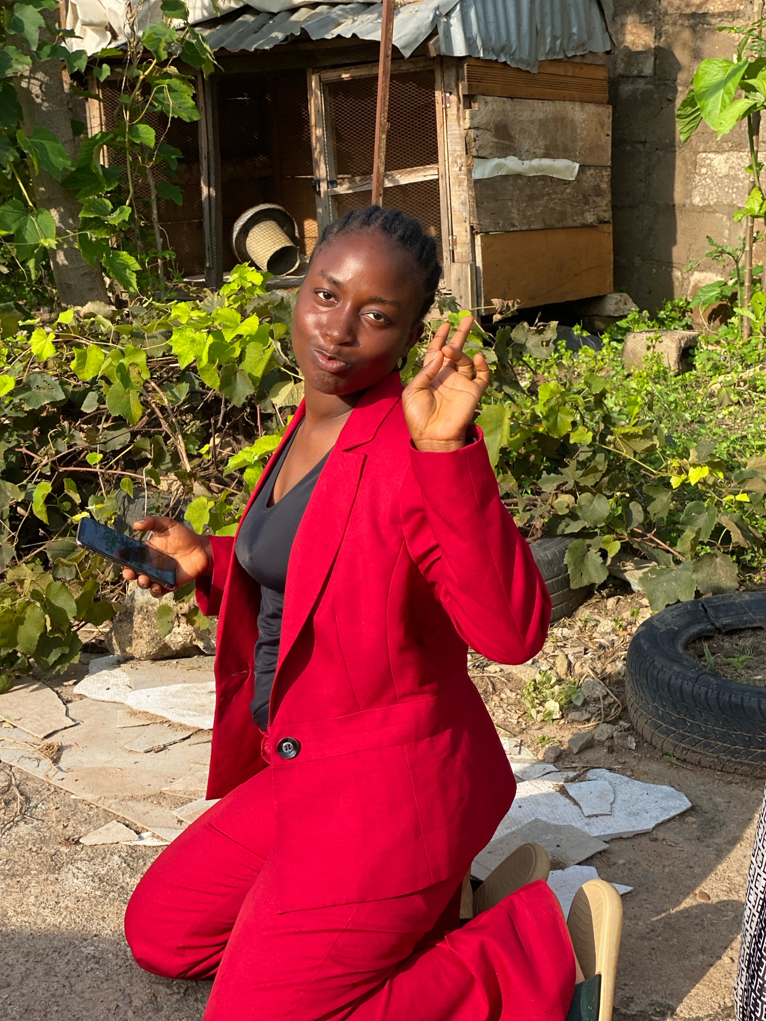 A young Black woman is kneeling outdoors, wearing a red blazer and matching pants. She is holding a black smartphone in her left hand and making a peace sign with her right hand. The background shows a rustic building with a corrugated metal roof, overgrown vegetation, and a discarded tire. The ground is uneven and covered with dirt and broken concrete. The overall scene suggests an informal setting, possibly a backyard or alleyway, in Sabon Tasha, Nigeria.