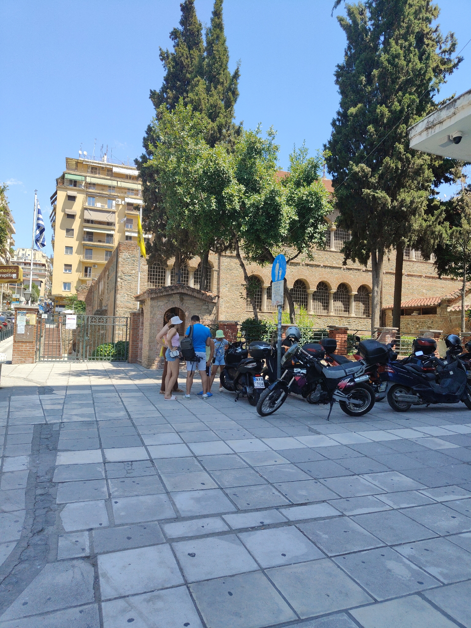 This is a street-level, slightly angled, medium-shot photograph taken in Thessaloniki, Greece, likely on a sunny day given the shadows. The foreground is dominated by a paved sidewalk made of square grey tiles, some of which appear worn or cracked. To the left of the sidewalk, there's a narrow strip of darker material, possibly a gap or a different type of paving.

In the middle ground, a group of four individuals, appearing to be tourists, stand on the sidewalk. Two women, dressed in casual summer clothing, are on the left. One wears a white hat, a pink tank top, and denim shorts. The other, further back, is not fully visible. Two men are on the right; one wears a blue t-shirt and white shorts, carrying a backpack, and the other wears a light-colored t-shirt and shorts. They appear to be conversing.

Behind the people, a metal fence with decorative elements encloses the entrance to a historical brick building with arched windows. This building, likely a church or an ancient monument, features red-tiled roofing and stone walls. A Greek flag (blue and white) and a yellow flag are visible on flagpoles near the building.

To the right of the building, several motorcycles and scooters are parked along the sidewalk. They are of various models and colors, including black and dark blue. One scooter is parked closest to the right edge of the frame.

In the background, a tall, pale yellow apartment building stands out, with multiple balconies and awnings. Several trees with dense green foliage are scattered throughout the scene, framing the building and the sky, which is clear and blue. A street sign with a blue pedestrian symbol is also visible near the parked motorcycles. The overall impression is of a busy urban environment with historical architecture and a lively street scene.