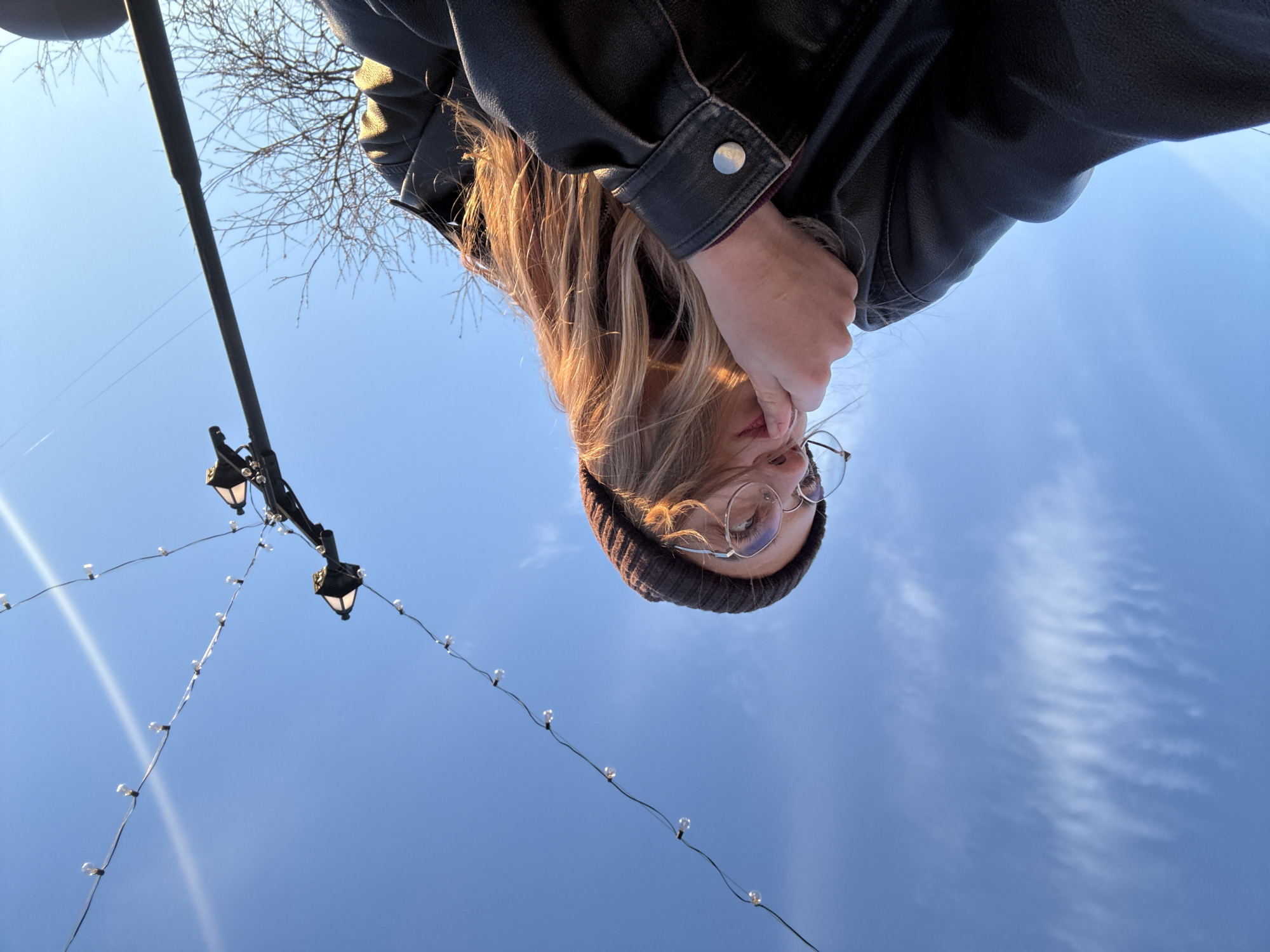 A low-angle outdoor shot captures a young person from below, appearing upside down against a clear blue sky. The person has long blonde hair, which is illuminated by sunlight, and wears a dark beanie, round glasses, and a black leather jacket. Their face is partially obscured by their hand.
In the upper left background, the intricate, bare branches of a tree are visible, suggesting a colder season like late autumn or winter. A tall, dark pole or lamppost runs vertically on the left side of the frame, from which several strands of decorative string lights with small clear bulbs are strung across the sky. Two antique-style street lamps are also suspended, contributing to an urban or park setting.
The sky is bright blue with faint, wispy clouds, indicating a clear, sunny day. The unique perspective suggests the person is looking down towards the camera while posing for a photograph. No text is visible in the image.