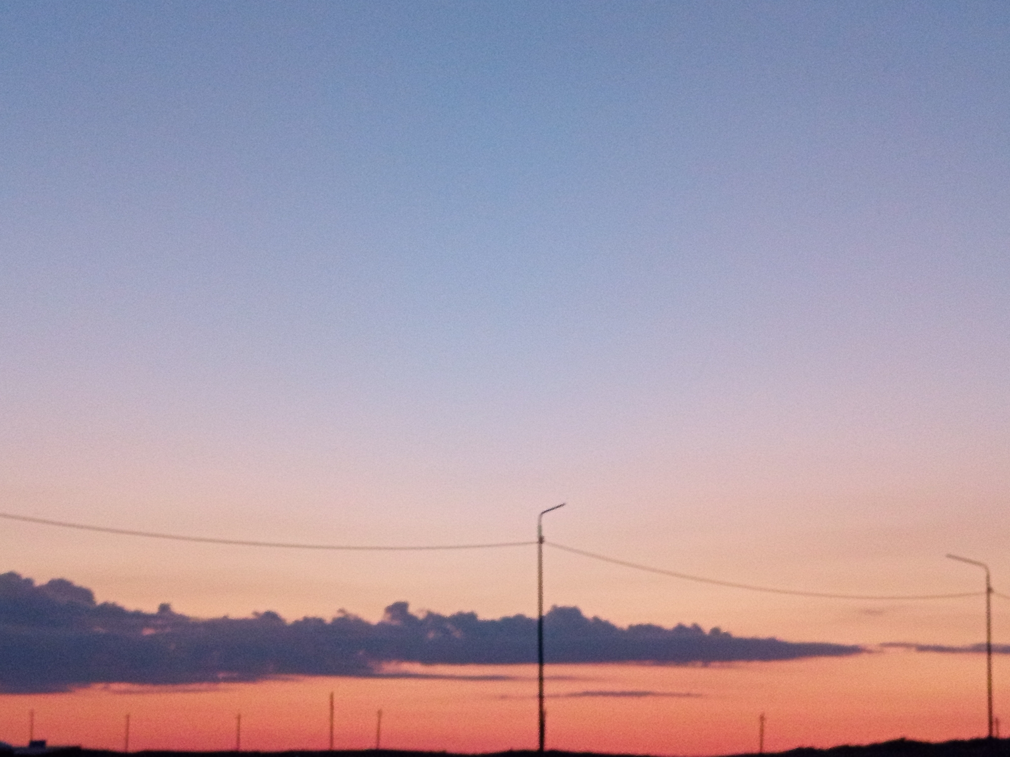 The image depicts a twilight sky over a flat, indistinct horizon line, likely in or near Баянауыл ауылдық округі, Kazakhstan. The sky transitions from a pale blue at the top to hues of pink and orange near the horizon. A dark, low-lying cloud formation is visible across the lower third of the sky. A thin, horizontal wire stretches across the frame, supported by an unseen structure. Two streetlights, silhouetted against the colorful sky, are present: one on the right with a visible curved arm, and another partially visible on the far right edge of the frame. Several thin, vertical poles or posts are also visible along the horizon line. The overall impression is of an open, somewhat rural or roadside landscape at dusk.