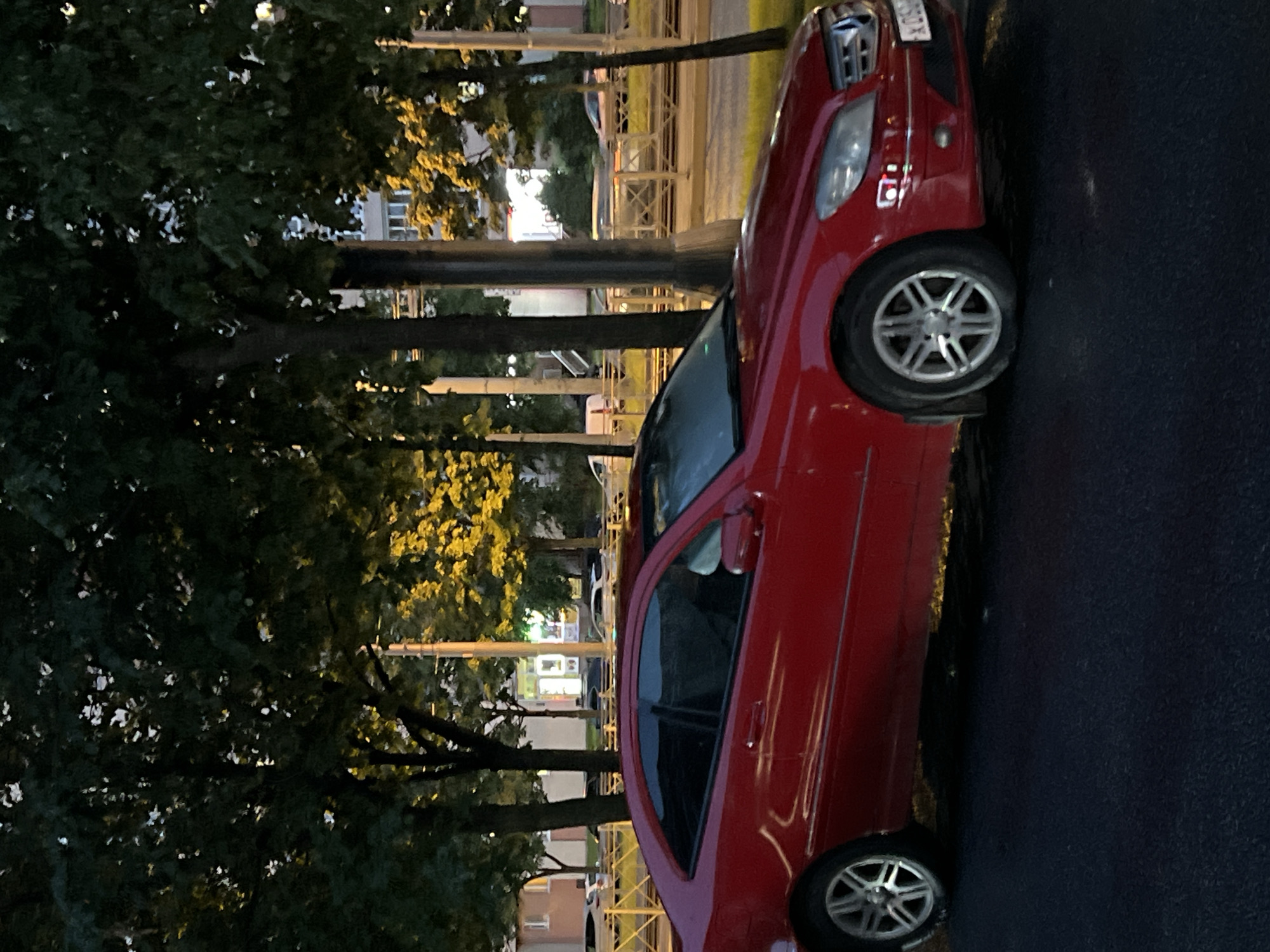 The media file is a slightly tilted, vertical, full shot of a red car parked on an asphalt road. The car is the primary subject. It is a two-door coupe with tinted windows and silver alloy wheels. A license plate is visible on the rear of the car, with some text and numbers. The car is parked next to a sidewalk and a grassy area with trees. The trees are tall and leafy, with some yellowing leaves, suggesting it might be autumn. The lighting is dim, with yellow streetlights or lamps casting a warm glow on the scene, indicating it is likely evening or night. In the background, buildings and streetlights are visible, suggesting an urban or suburban setting. The scene appears to be an ordinary street in a residential or commercial area. There is no discernible activity or event taking place; the car is simply parked. The overall mood is calm and quiet, typical of a street at dusk or night. The visible text on the license plate appears to be "X 095 OX".