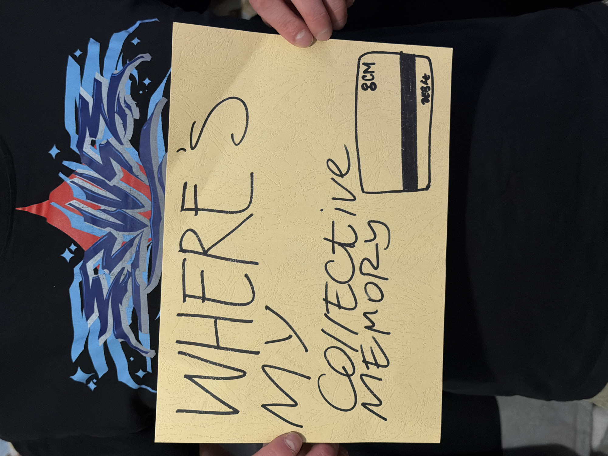 A person wearing a black t-shirt with a graphic design in blue, silver, and red is holding up a yellow card. Written on the card in black marker are the words "WHERE'S MY COLLECTIVE MEMORY". A drawing of a credit card with "8CM" and "2024" is also on the card. The location of the photo is not evident.