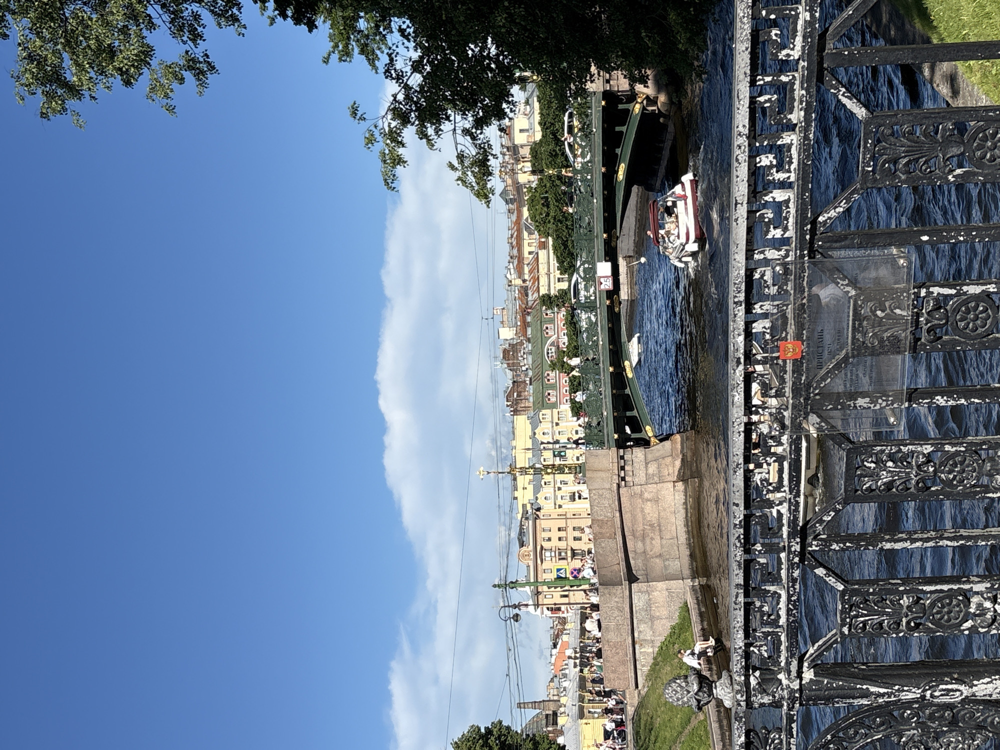 This image captures a vibrant daytime scene along the Moika River in Saint Petersburg, Russia, likely from an embankment or a bridge near the Pevchesky Bridge, looking eastward.
The foreground features a weathered, black cast-iron railing with an intricate classical pattern, showing visible chips and peeling white paint. Attached to the railing is a small, rectangular red emblem, possibly a Russian state symbol, and a translucent plaque with faded Russian text, including the word "ПРИСТАНЬ" (PIER or LANDING STAGE).
Beyond the railing, the Moika River flows, its surface agitated with ripples and reflecting the bright sunlight in dark blue hues. A white and red motorboat with a dark canopy is visible in the mid-river, traveling from right to left, with several individuals aboard. Further upstream, a smaller, lighter-colored boat or object is partially obscured by the bridge structure.
The midground is dominated by a multi-arched bridge, painted dark green with ornate metal railings and supporting stone abutments on both banks. Numerous pedestrians are visible walking on the bridge, and several vehicles, including white and yellow taxis, are present. A white square sign with the number "2.5" in red is attached to the bridge structure. On the left bank, below the bridge, a grassy slope descends to the water's edge, where a stone ledge provides seating for a few individuals. One person is seated directly on the ledge near the water, and another on a stone ornament resembling a pineapple or pinecone.
The background consists of a dense urban landscape characterized by rows of multi-story buildings lining both sides of the river. These structures display varied architectural styles and a palette of pastel colors including yellow, orange, green, blue, pink, and white. Overhead power lines and streetlights crisscross the scene above the buildings. Green trees with lush foliage frame the image on both the left and right sides, with significant tree cover on the right bank. The sky above is a clear, bright blue with scattered, fluffy white cumulus clouds.