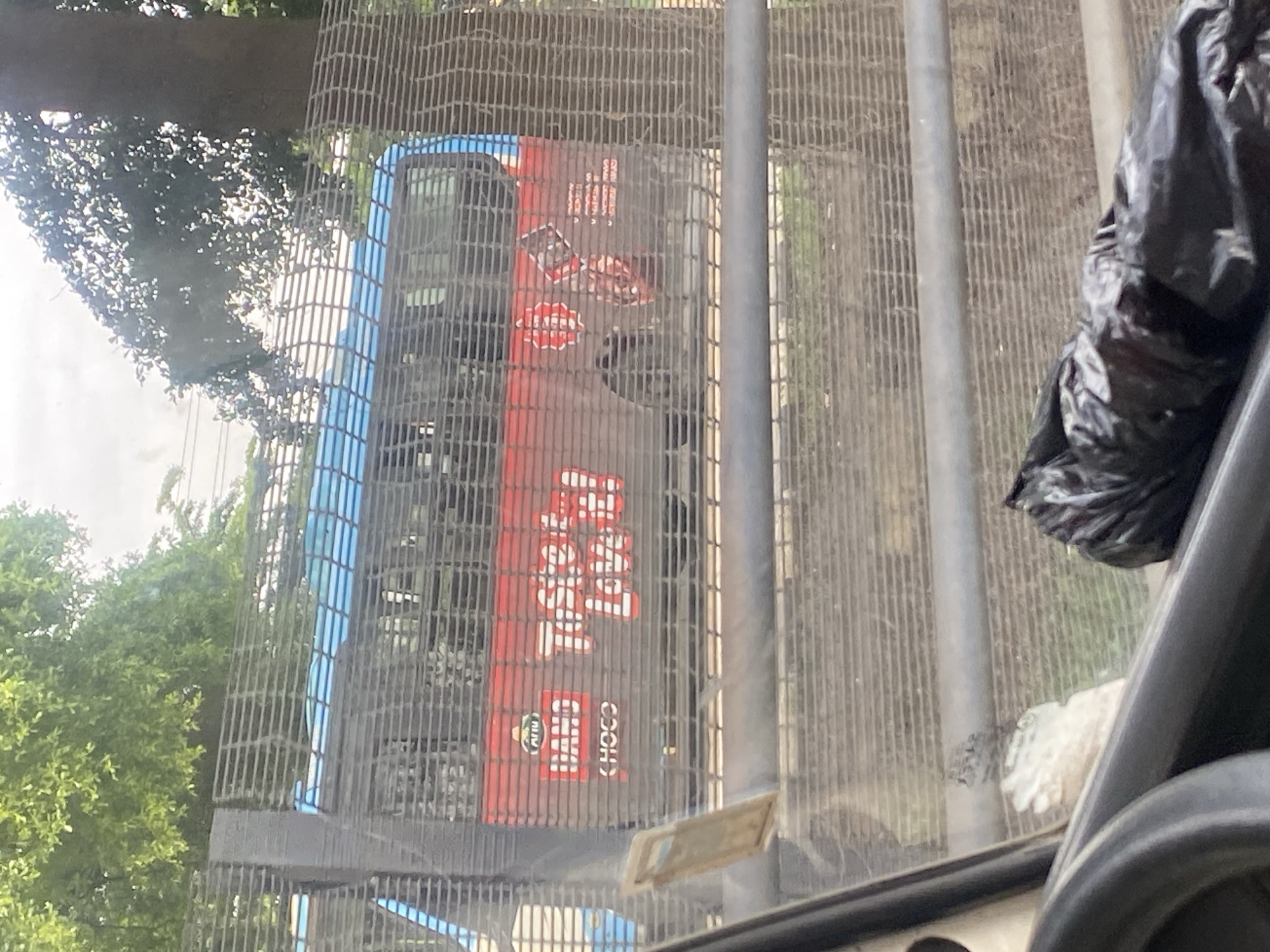 An outdoor scene captured from inside a vehicle, looking through a fence at a parked bus. The bus is predominantly blue and red, featuring the text "Taste the LOVE!" and "HAND CHOGO" prominently displayed on its side. Several circular emblems are also visible on the bus. The fence, constructed from wire mesh, runs horizontally across the frame. Beyond the fence, trees and a cloudy sky are visible. Inside the vehicle, a portion of the steering wheel and a black plastic bag are in the foreground. The location is specified as Ikorodu, Nigeria.
