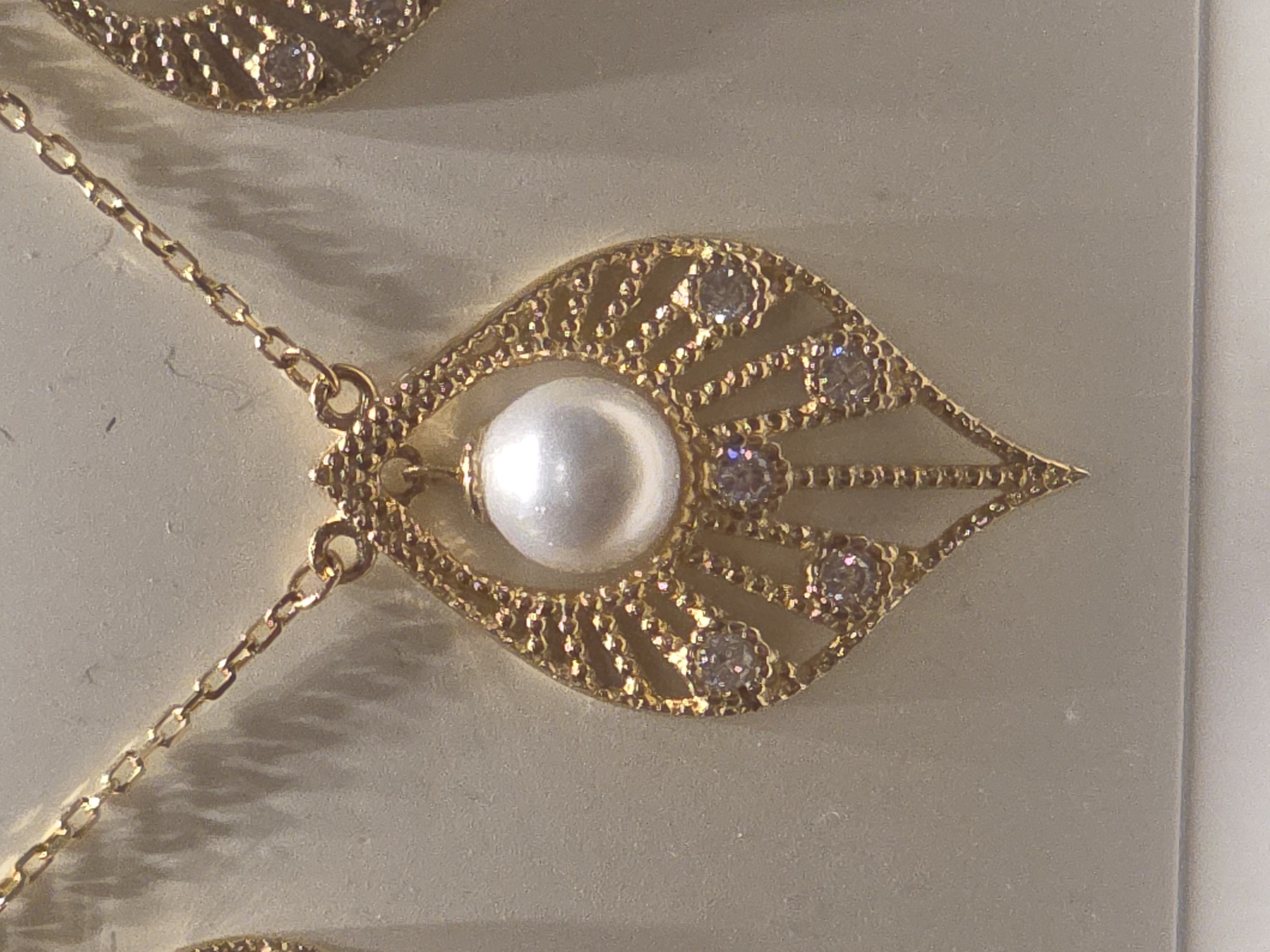 A close-up image shows a gold-colored necklace with a pearl and diamond-like gems, resting on a white or light-colored surface. The necklace features a delicate chain and an ornate pendant in a leaf shape, with the pearl positioned in the center. Diamond-like gems are distributed around the pearl and along the edges of the pendant. The lighting suggests the image was taken indoors. There is no indication of the location of Warsaw, Poland.