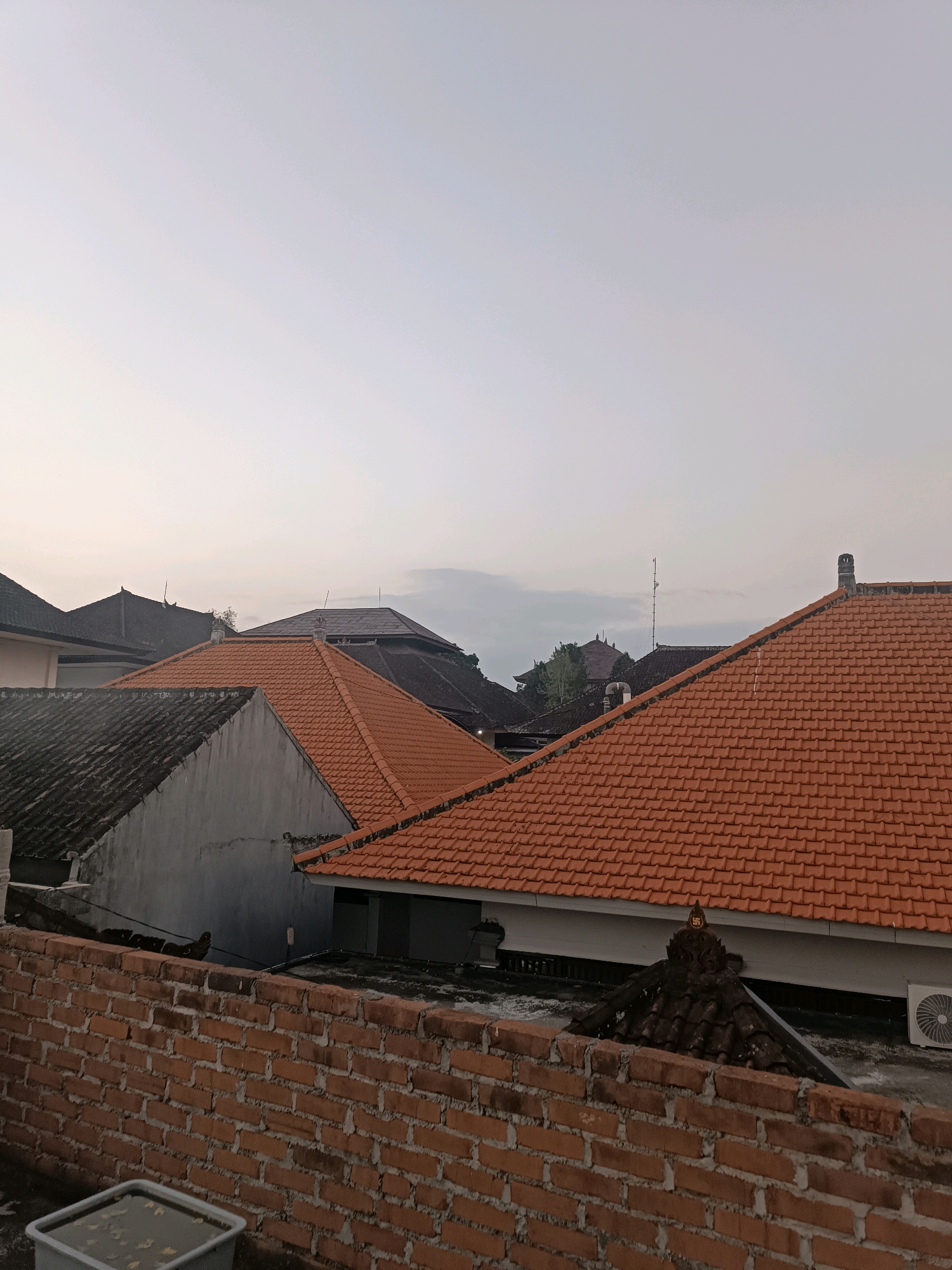 This image depicts a view of rooftops in Tabanan, Indonesia, under a pale, overcast sky. The foreground features a partially visible brick wall. Beyond the wall, several tiled rooftops are visible, dominated by red clay tiles and some darker, older tiles. Architectural elements include pointed roof peaks and what appears to be a small shrine or decorative element on one of the darker roofs, featuring a symbol that resembles a swastika. Several buildings with various roof styles are clustered together, with trees visible in the background. An air conditioning unit is mounted on the side of one of the buildings. The overall impression is an urban or semi-urban residential scene.
