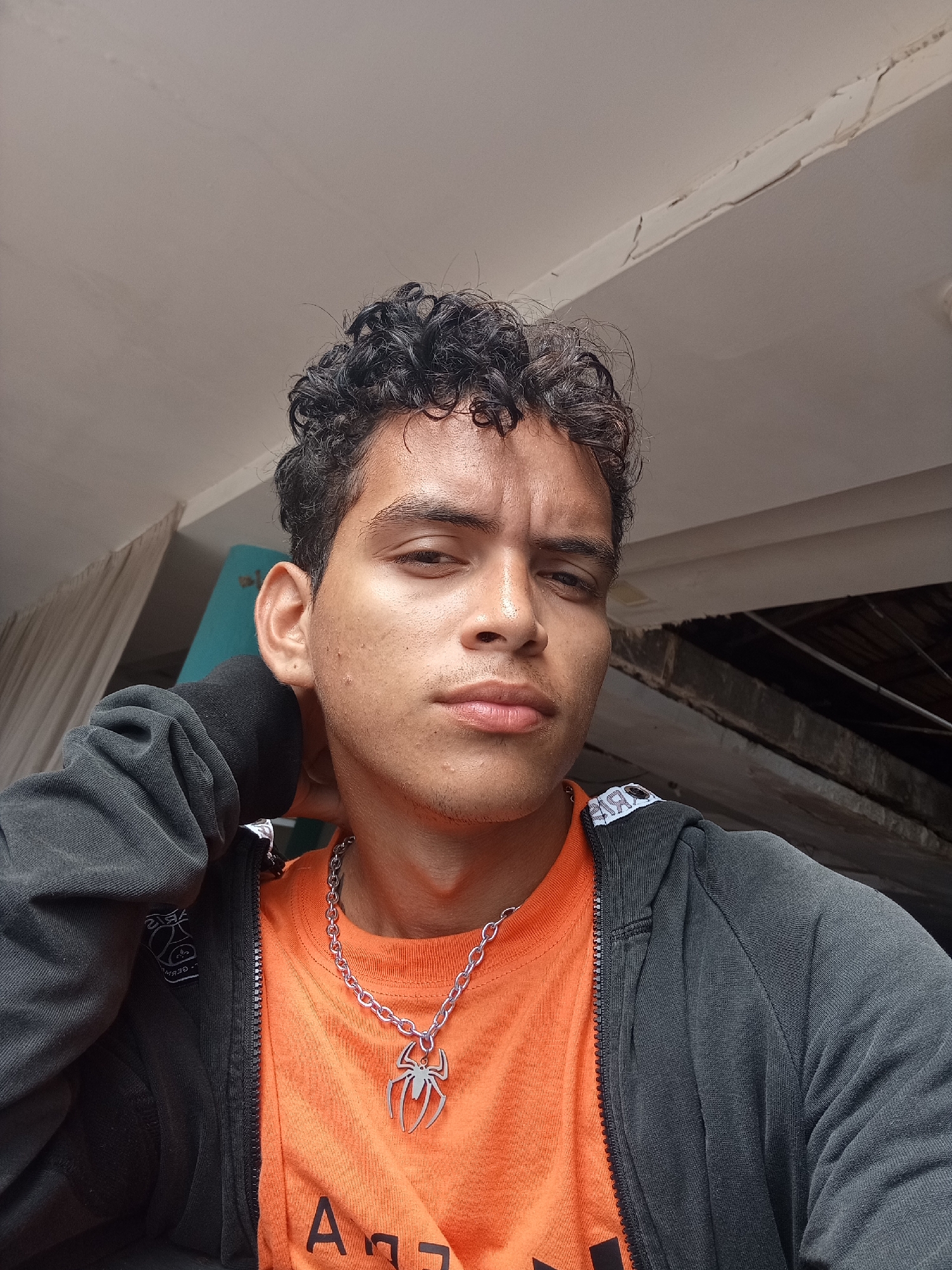 A medium shot shows a young man with curly dark hair, wearing an orange t-shirt with text, a dark hooded zip-up jacket, and a silver chain necklace with a spider pendant. He is holding his right hand behind his neck. The background includes a white ceiling with cracking paint, a blue and white surface, and dark, indistinct structures. The photo was taken in Playa El Agua, Venezuela.