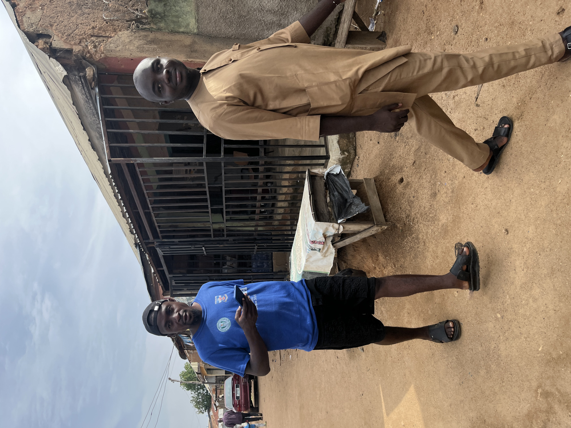 In Jos, Nigeria, two men walk down a dirt road lined with buildings. The man on the left wears a blue t-shirt, black shorts, and sandals, holding a smartphone in his hand. The man on the right is dressed in a tan, traditional outfit and sandals, walking slightly ahead. In the background, a red car is parked, and various structures with corrugated metal roofs and barred windows suggest a market or commercial area. The ground is composed of reddish-brown earth, and the sky is overcast.