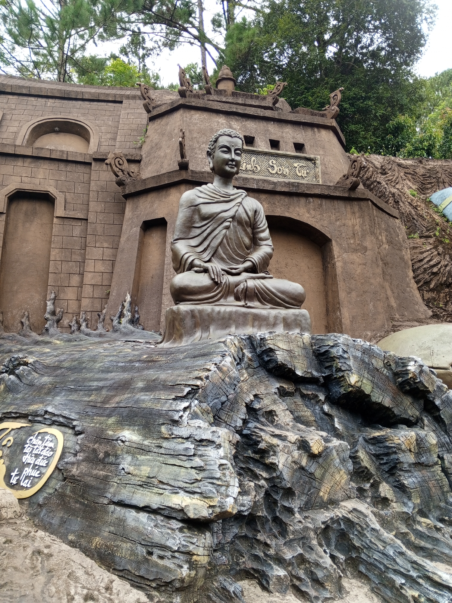 A statue of Buddha in a seated meditation pose is positioned on a rough, textured base made of what appears to be carved wood or rock. The statue is made of a dark material, likely bronze or a similar metal, and depicts Buddha with serene facial features and draped robes.
Behind the statue is a large, textured wall, possibly part of a temple or monument. The wall has architectural details, including recessed arched doorways and decorative elements along the top. There is a plaque with text on the wall above and to the right of the Buddha statue.
The environment appears to be outdoors, with lush green trees visible in the background and a cloudy sky overhead. In the foreground, to the left of the Buddha statue, there is a circular plaque with ornate gold lettering and a white border, also set into the textured base.
This scene is located at Thien Vuong Pagoda in Da Lat, Vietnam. The pagoda is known for its impressive architecture and beautiful natural surroundings.