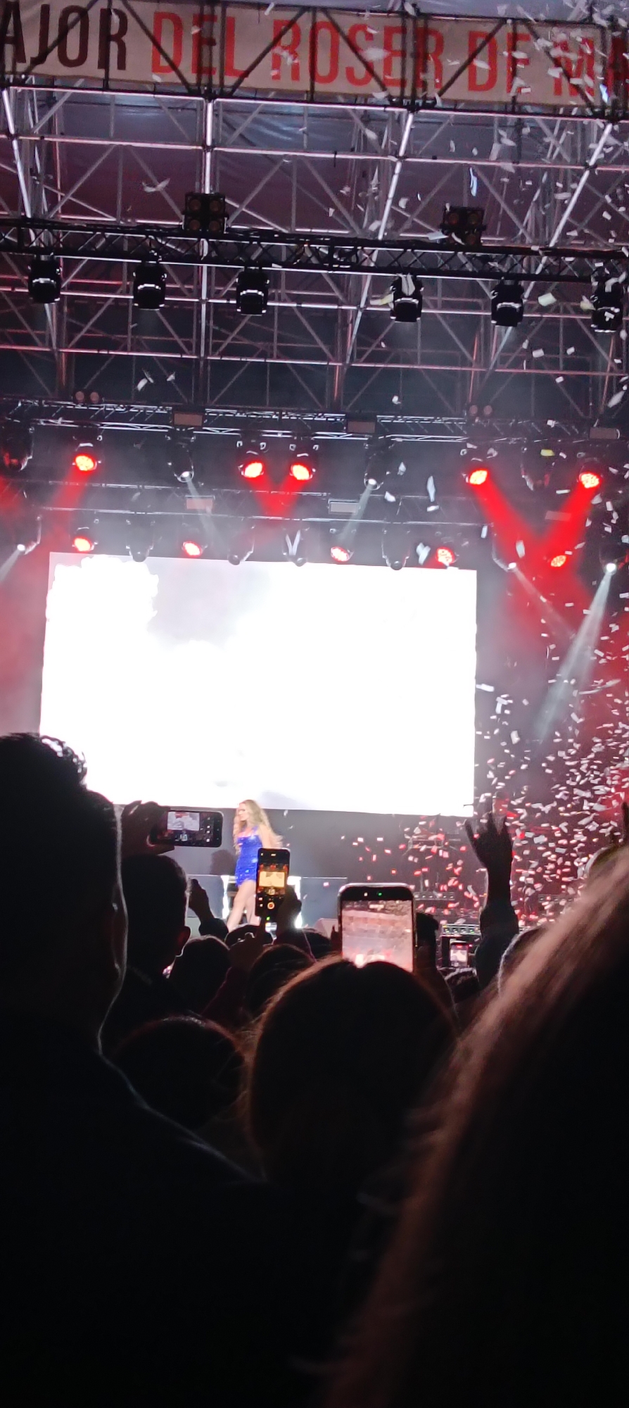 The image captures a concert scene in Cerdanyola del Vallès, Spain. A female singer in a short blue dress is on stage, illuminated by bright white stage lights and red spotlights. Confetti is falling around the stage. The audience is visible in the foreground, holding up phones to record or photograph the performance. A large white screen dominates the back of the stage. Above the screen, there is a metal structure with stage lights. A banner that says "AJOR DEL ROSER DE SO" is visible above the metal structure.
