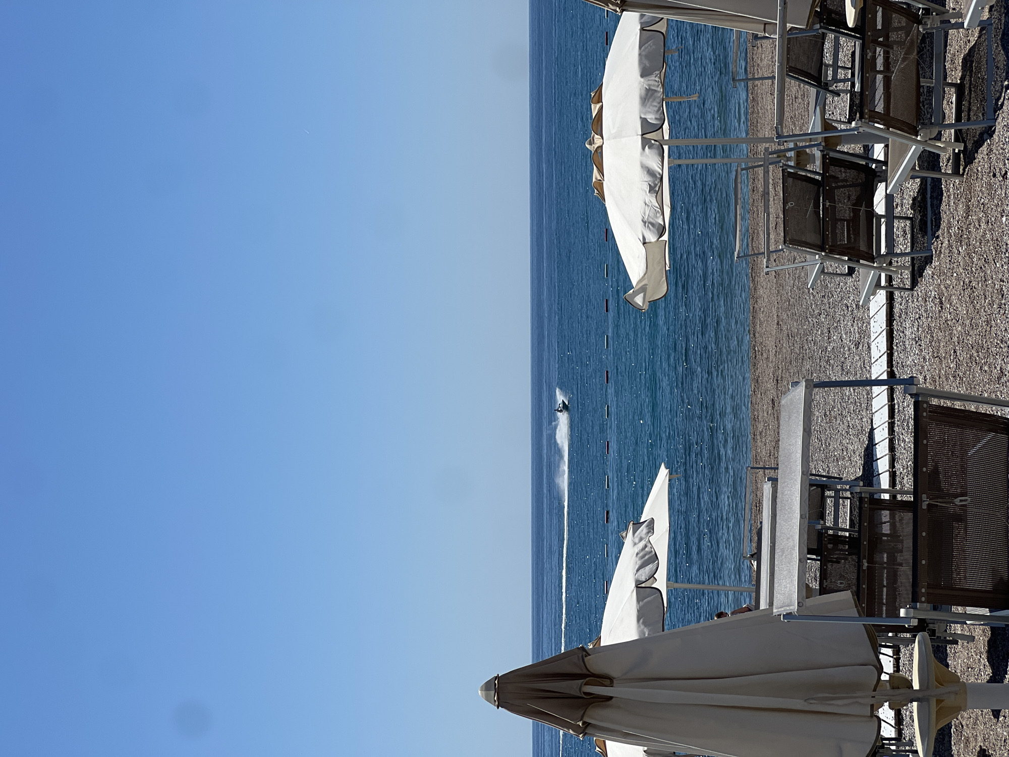 The image captures a coastal scene near Prijevor, Montenegro, featuring a beach with sun umbrellas and lounge chairs. The Adriatic Sea stretches out towards the horizon, with a personal watercraft leaving a wake. The sky is a clear blue. The beach appears to be pebbly.