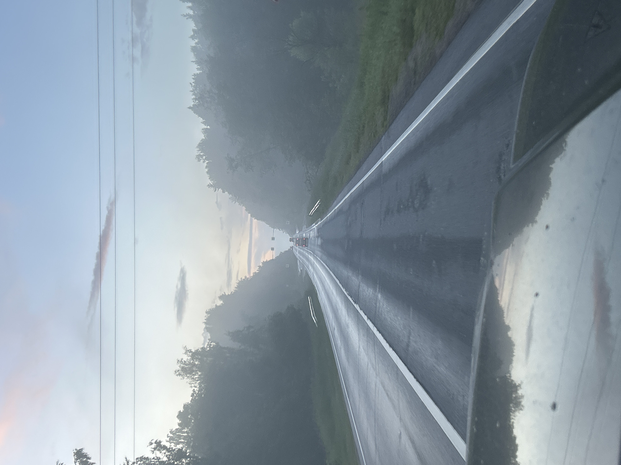The image shows a wet, two-lane asphalt road in Russia, likely during a foggy or misty morning or evening. The road is bordered by dense, dark green trees on both sides, which appear to be covered in fog or mist, obscuring the horizon. The sky is a pale blue with hints of pink from a setting or rising sun, and there are three power lines stretching across the upper part of the frame. The road surface reflects the light and the surrounding environment, indicating recent rain. Several vehicles are visible in the distance, traveling in the same direction as the camera. The foreground shows a portion of a dark-colored vehicle, likely a car, from which the photograph was taken, its hood also reflecting the sky. The overall atmosphere is serene and slightly melancholic due to the weather conditions and the vastness of the landscape.
