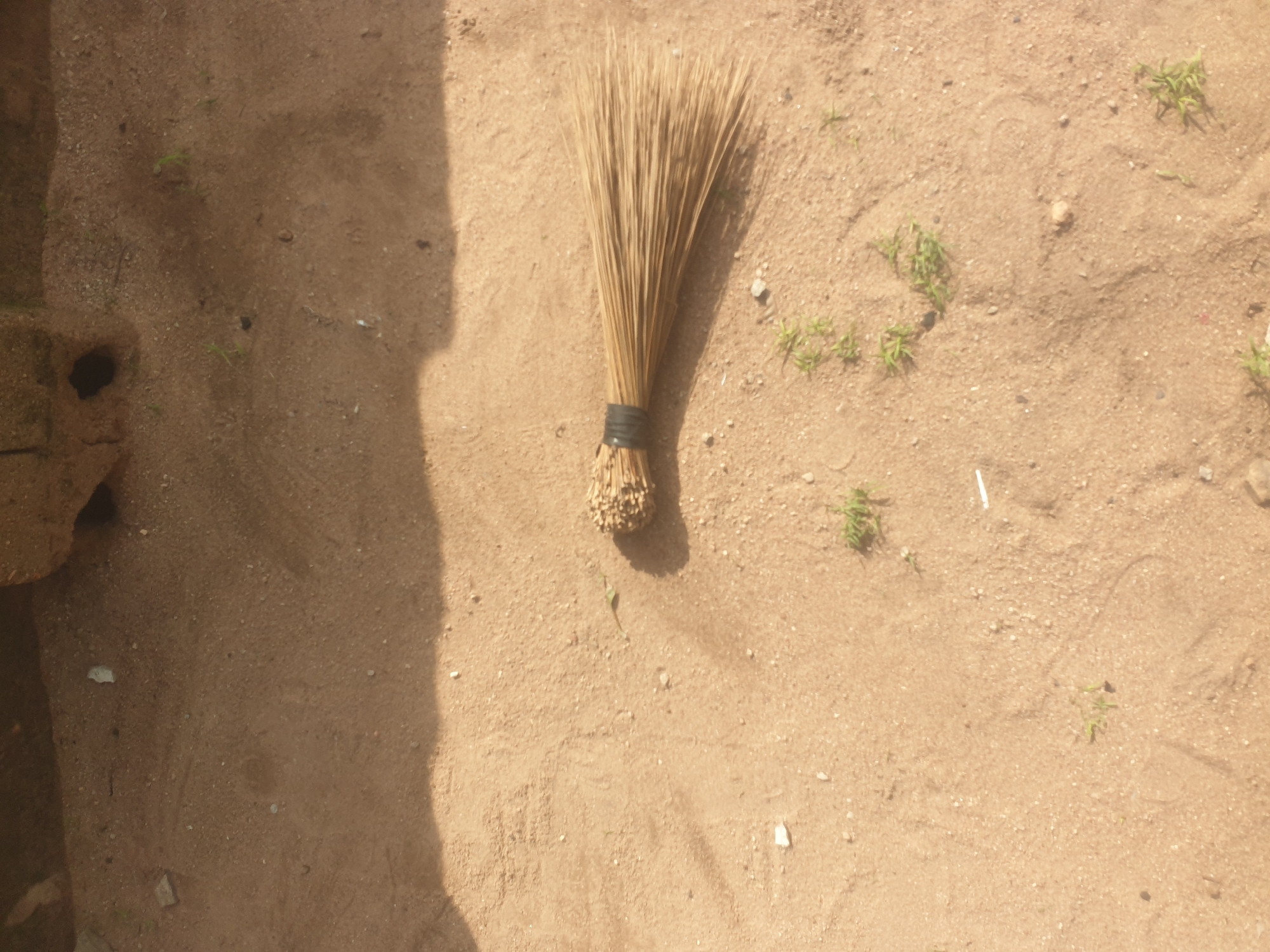A light brown broom made of twigs is lying on sandy ground, its bristles fanned out to the right and its handle tied with a black band. The ground is dry and dusty, with small pebbles and patches of sparse green vegetation visible. A shadow falls across the upper left portion of the image, indicating an overhead light source. The background appears to be a rough, textured wall made of bricks or mud. The scene is outdoors, possibly in a yard or pathway in Jauro Yinu, Nigeria. There are no people or animals present.