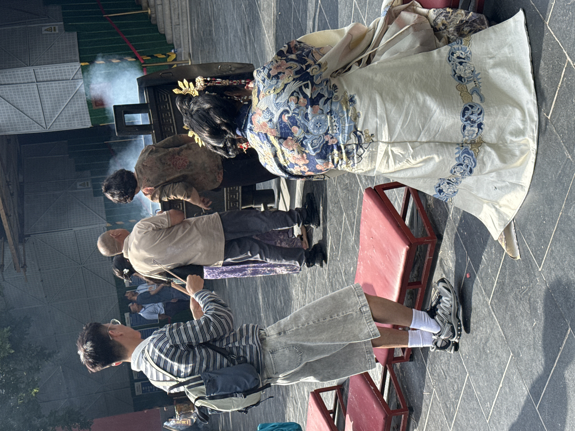 In a courtyard in Dongcheng District, China, on a paved surface with square tiles, a group of people are gathered. In the foreground, a person in a traditional ornate robe with intricate blue and white floral and bird patterns is kneeling on a red mat. Their elaborate headdress features gold ornaments and dangling embellishments. To the left, a young person wearing a striped long-sleeved shirt, light-colored culottes, white socks, and athletic shoes stands holding a wooden stick. They have a backpack on their shoulders. Behind them, other individuals are visible, including a person in a light-colored t-shirt and dark pants, and another in a patterned brown top and dark pants. A large, dark incense burner is present, emitting a plume of white smoke. In the background, a construction fence and scaffolding suggest ongoing work. Red ropes are strung across the scene, and stairs are visible to the right. The lighting indicates it is daytime, with sunlight casting shadows.