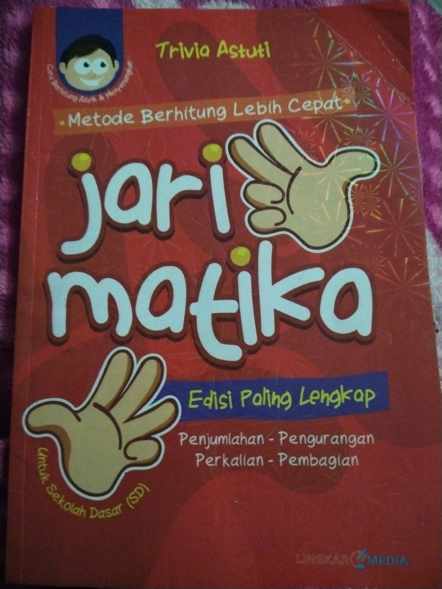 The image is a close-up of the cover of a book titled "Jari Matika" which translates to "Finger Math". The book is red with white and yellow lettering. The title "Jari Matika" is large and centered on the cover. Above the title, there is a smaller text that reads "Metode Berhitung Lebih Cepat", meaning "Faster Calculation Method". There is also a cartoon drawing of a child's head with a smiling face at the top left corner, and a cartoon drawing of an open hand with fingers extended at the bottom left. The hand drawing is surrounded by text that reads "Untuk Sekolah Dasar (SD)", meaning "For Elementary School (SD)". At the top of the book, the text "Trivia Astuti" is visible. Below the large title, a blue banner contains the text "Edisi Paling Lengkap", meaning "Most Complete Edition". Underneath this, the text "Penjumlahan - Pengurangan Perkalian - Pembagian" (Addition - Subtraction Multiplication - Division) is written. At the bottom right corner of the book cover, a logo with the text "LINGKAR MEDIA" is visible. The book cover has a textured background with starburst patterns. The image is taken from a slightly elevated angle, showing the book resting on a textured, possibly fabric surface, which appears to be a bedspread or blanket, with a purplish hue. The lighting is diffused, suggesting an indoor setting, possibly a bedroom or study. There are no people visible in the image, and the focus is solely on the book. The image quality is good, with clear text and illustrations.