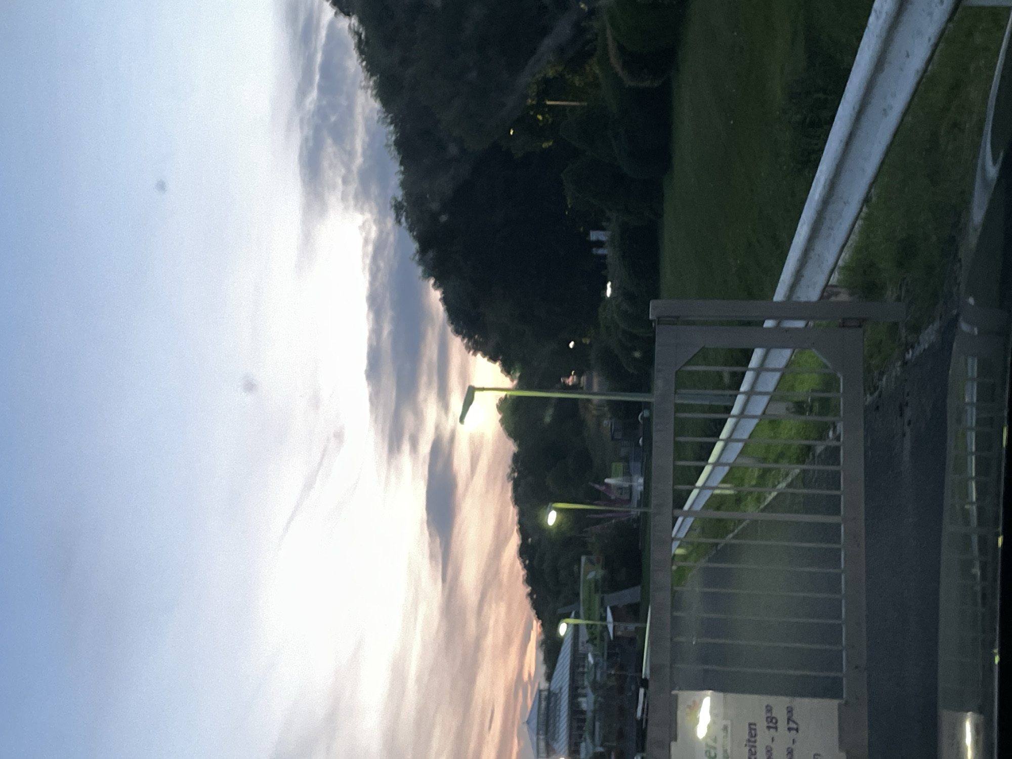 The image depicts a street scene at dusk in Siegburg, Germany. In the foreground, a metal gate with vertical bars is visible, separating the viewer from a paved area and a grassy slope. A white metal guardrail runs along the right side of the image, parallel to a road.
In the midground, several streetlights with green fixtures illuminate the scene. Behind them, a cluster of trees forms a dark, dense line against the twilight sky. A building with a modern, glass-paneled roof and a sloped design is visible on the left side of the midground, possibly a commercial establishment. A sign with German text, including "zeite", "00 - 18.30", and "00 - 17.00", is attached to the gatepost on the left.
The sky is a gradient of blues and purples, with soft, wispy clouds tinged with orange and pink from the setting sun. The overall atmosphere is calm and serene. No people are visible in the image.