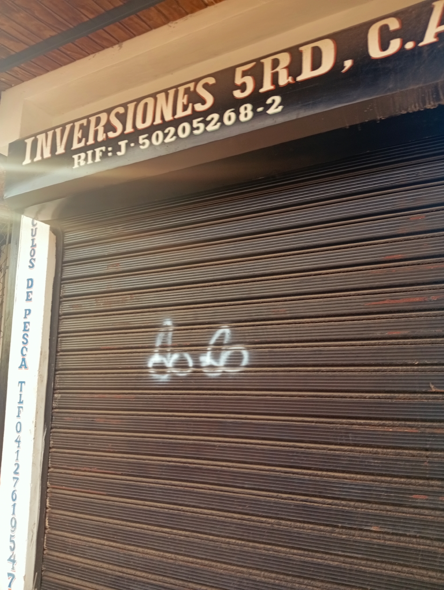 The image shows the exterior of a business with a dark brown metal roll-up door. Above the door, a sign reads "INVERSIONES 5RD, C.A." with the RIF number "J-50205268-2". To the left of the roll-up door, a white vertical sign lists "CULOS DE PESCA" followed by a phone number "TLF 04127619547". There is graffiti in white spray paint on the roll-up door, appearing to be two stylized representations of male genitalia. The overall impression is that of a commercial establishment in an urban or semi-urban setting in Juan Griego, Venezuela.
