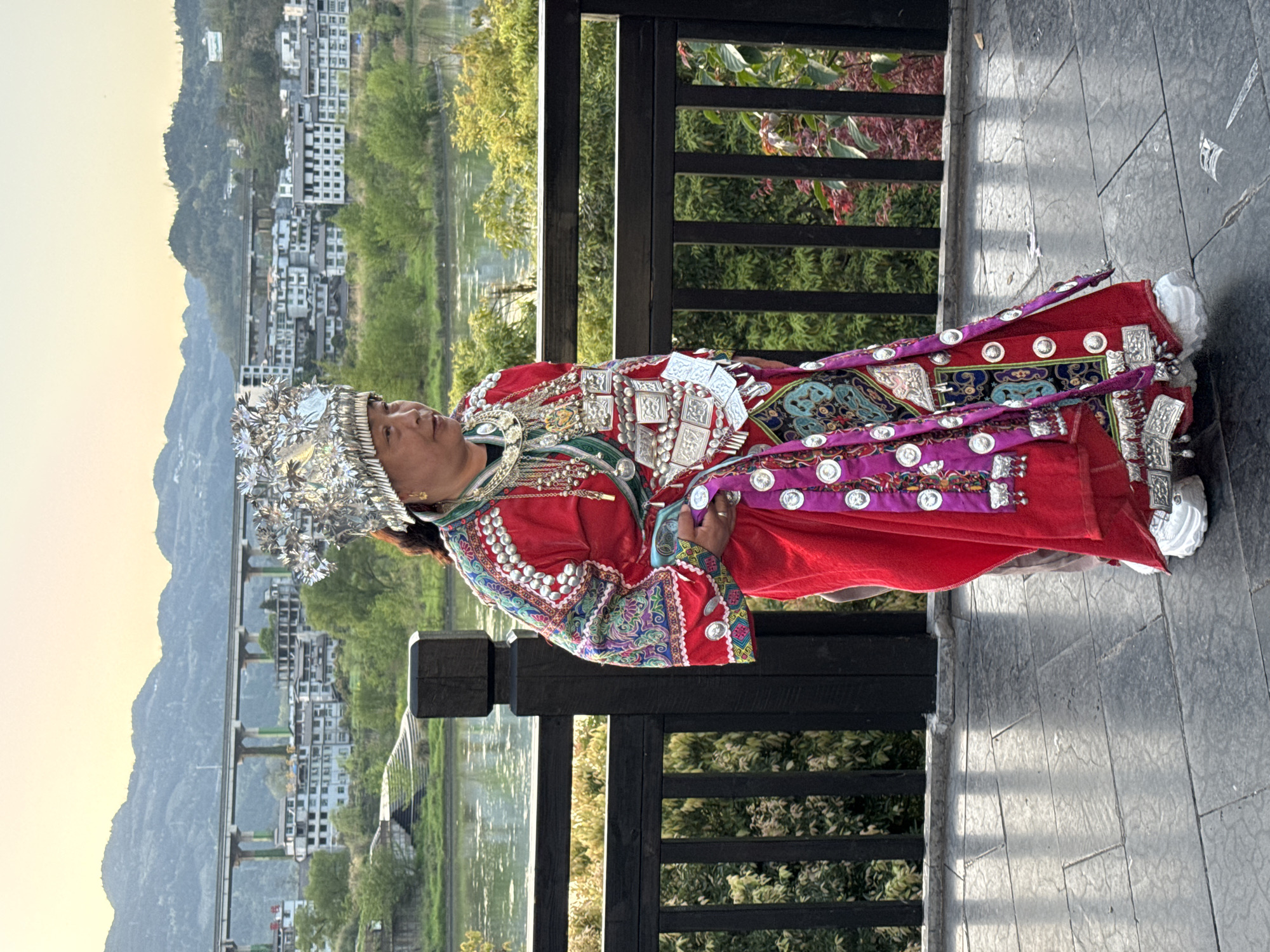 The image depicts a person wearing traditional Miao clothing in Zhuoshui, China. The individual is adorned with a large, silver headdress and a red garment embellished with silver ornaments, purple accents, and colorful embroidery. They are leaning against a dark wooden railing. The background features a town with buildings nestled into the hillside, a river or lake reflecting the sky, and distant mountains. The time of day seems to be late afternoon or early evening.