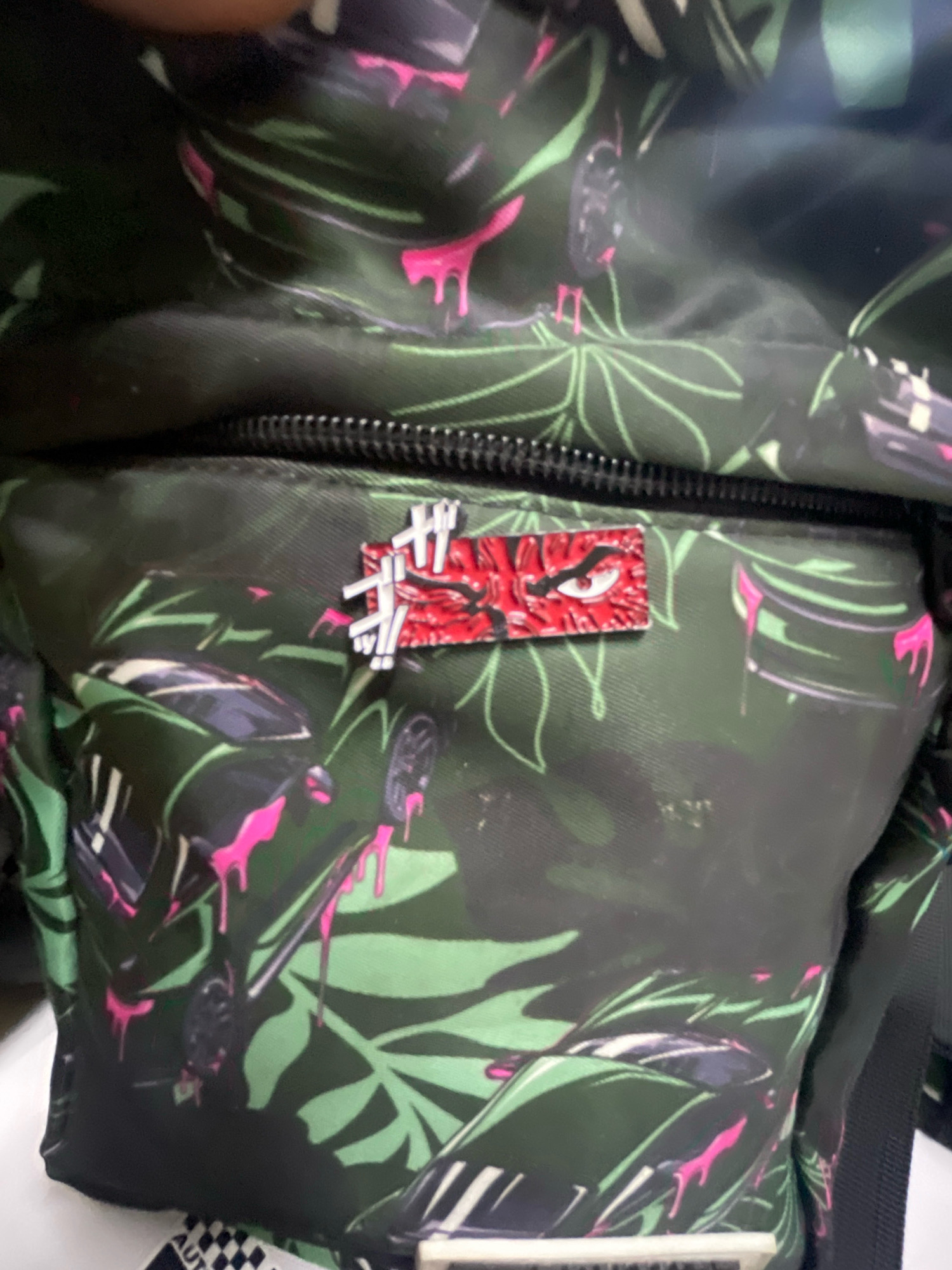 The image shows a close-up of a green backpack with a tropical leaf and car pattern. A red and white pin depicting a stylized eye is attached to the front zipper area of the backpack. The pin is rectangular with a red background and a white outline of an eye with sharp features and a fierce expression. The backpack's pattern includes large green leaves and elements that resemble car parts or reflections, with pink drips or accents throughout. The lighting suggests the photo was taken indoors, likely with a flash. The specific location in the city of Unknown, Unknown cannot be determined from the provided image.