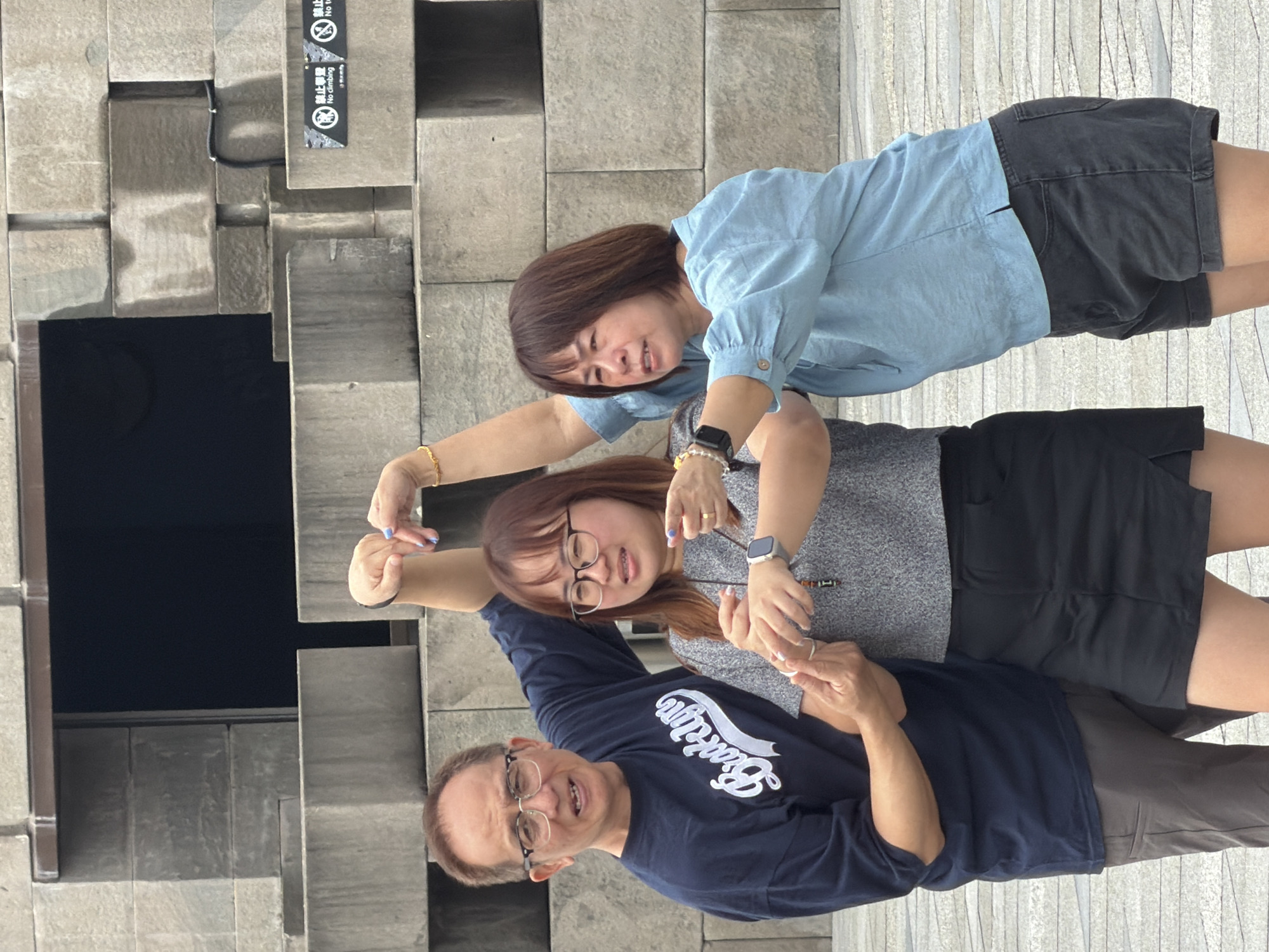 Three people are posing for a photo in front of a modern concrete structure. On the left, a man with glasses and a navy blue t-shirt smiles at the camera. In the center, a woman with glasses and a grey top holds hands with the man and points outwards. On the right, another woman in a blue shirt and dark shorts also points outwards. The background consists of large, interlocking concrete blocks, some of which have signs indicating "No climbing." The lighting suggests an overcast day.