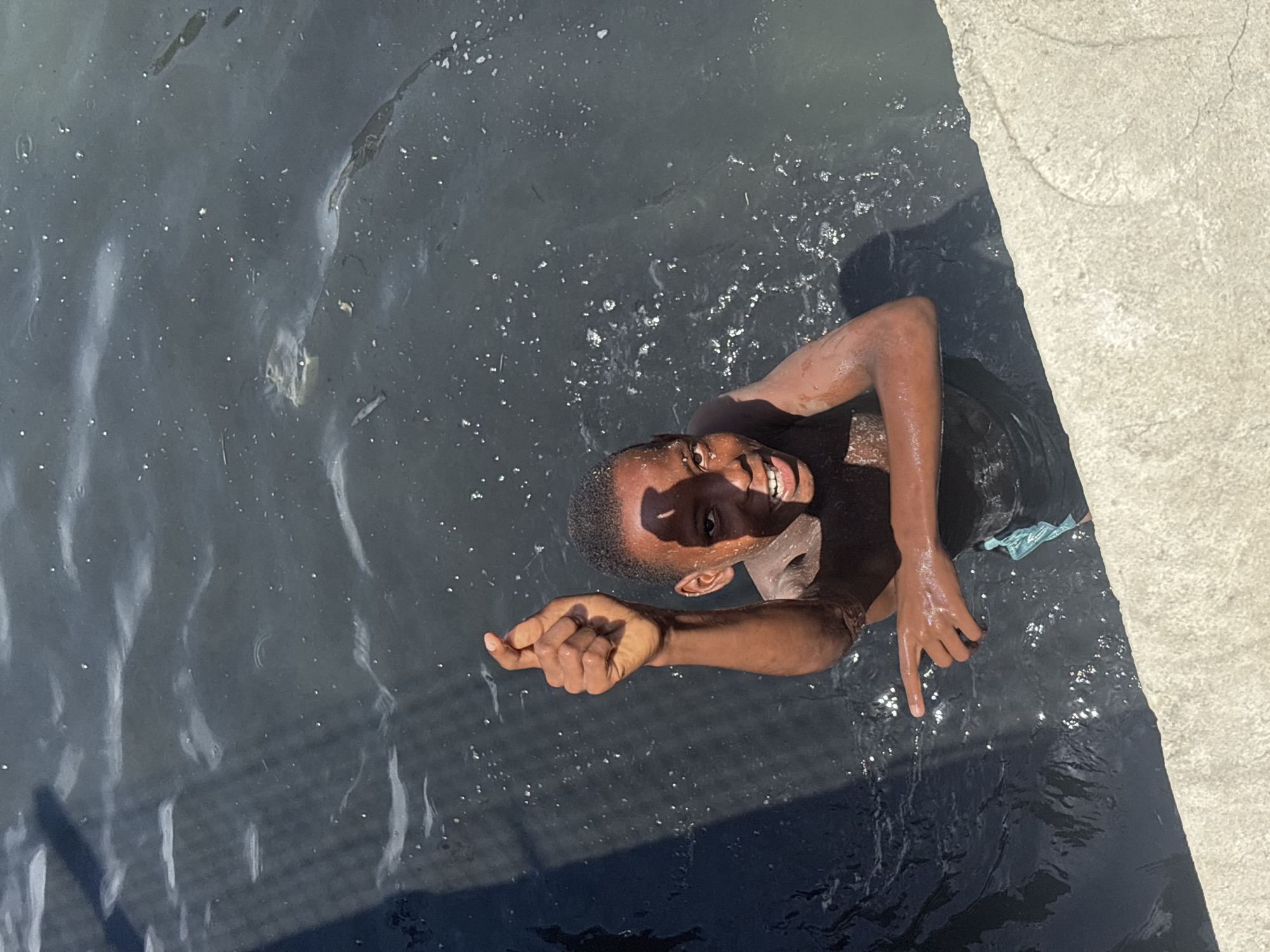 From an overhead perspective, a young Black boy is partially submerged in dark, murky water, looking up at the camera with a wide, joyful smile that reveals his teeth. His skin is wet and glistens under direct sunlight, which also creates bright reflections and speckles on the water's surface. His right arm is extended out of the water, with his hand curled and his index finger pointing towards the viewer. His left arm is also visible, with a glimpse of light blue-green fabric near his side, possibly part of his clothing.
To the right side of the image, a light-colored, textured concrete slab or edge of a structure is visible, suggesting the water body is man-made, possibly a canal or drainage ditch in Igboefon, Nigeria. The water contains small pieces of floating debris. The strong shadows and bright highlights indicate it is daytime with clear, sunny weather. The boy's expression conveys happiness and playfulness. No text is visible in the image.