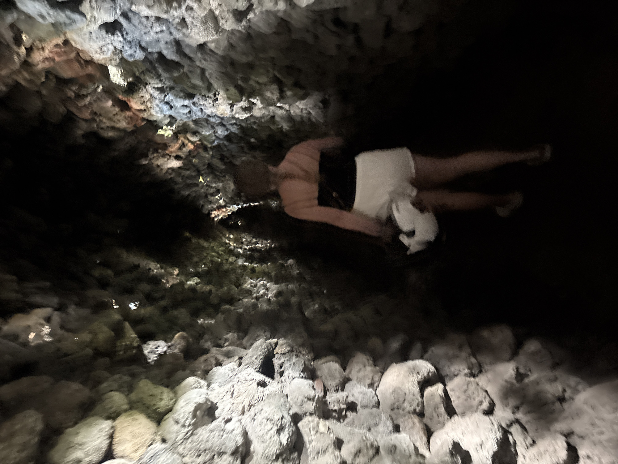 This image captures a person floating on their back in a dimly lit cave, likely located in Cam Ranh, Vietnam. The scene is dominated by rough, irregular rock formations that constitute the cave's walls and ceiling, illuminated by unseen artificial light sources that cast highlights and shadows across the textured surfaces. Some rock areas exhibit natural variations in color, including grays, whites, and reddish-brown hues.
The central subject is a single individual, seen from a high angle, lying horizontally in calm water. They appear to be wearing dark swimwear on their upper body and light-colored shorts or a towel wrapped around their lower body. Their face is partially obscured or indistinct, but their posture suggests a relaxed state of floating or gentle swimming.
The water surface in the foreground shows reflections of the rocky environment and light sources. The cave floor visible through the water and along the edges is also composed of similarly textured rocks. The setting is clearly an enclosed, underground environment, indicated by the lack of natural light and the characteristic cave structures. There are no discernible emotions or visible text in the image.