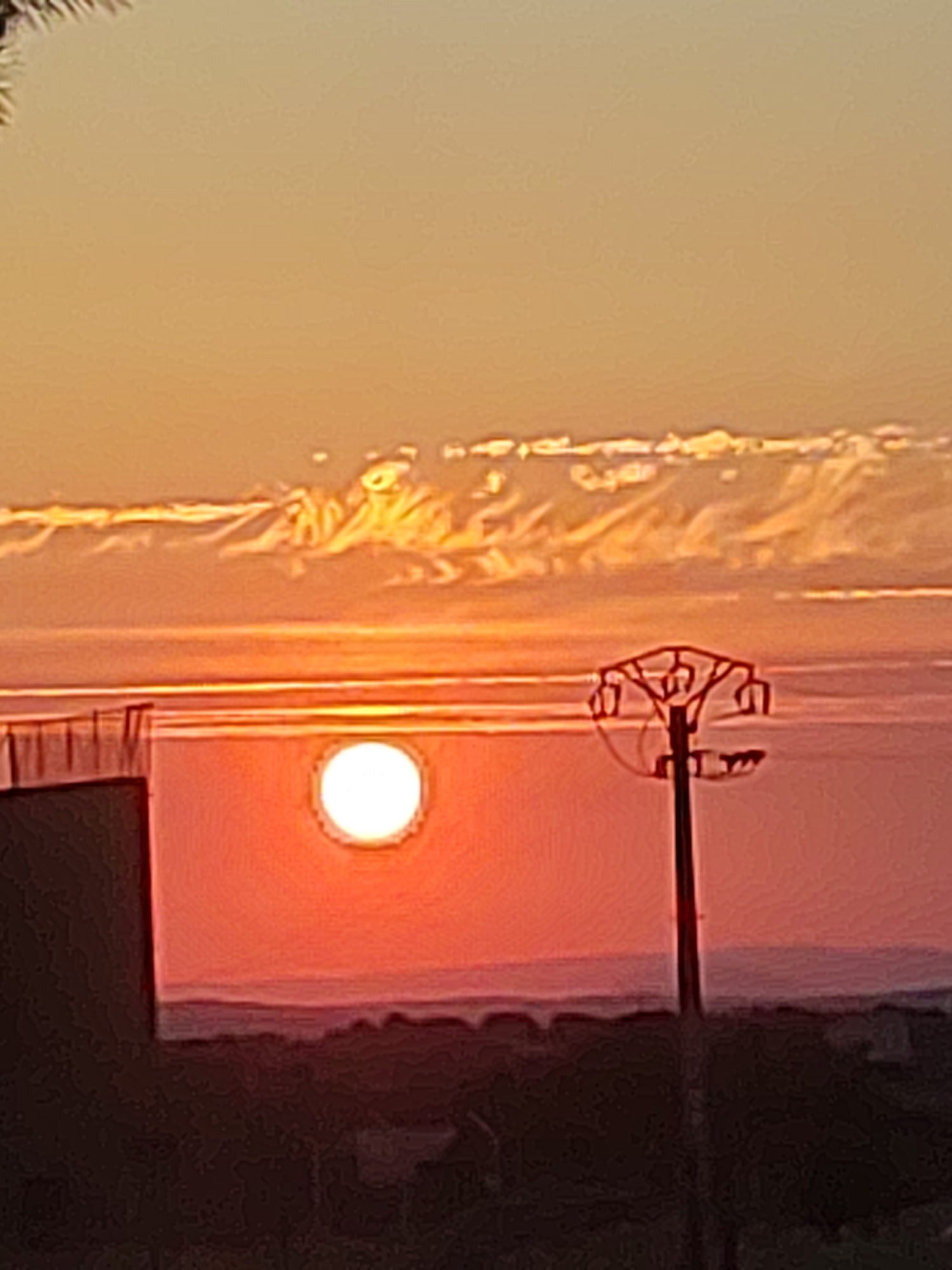 This image captures a vibrant sunset over a landscape, with the sun appearing as a bright, glowing orb. The sky transitions from a pale orange at the top to a deep red and pink hue closer to the horizon, suggesting the final moments of daylight. Wispy clouds are visible, illuminated by the setting sun, creating a dramatic effect.
In the foreground, a dark silhouette of landforms suggests hills or buildings. A prominent object, a power line pylon with insulators, stands starkly against the colorful sky. To the left, a building with a railing or fence is partially visible, and a circular window or opening on its facade prominently displays the sun. The overall scene evokes a sense of tranquility and the natural beauty of a sunset. The time of day is clearly evening, and the weather appears clear, allowing for an unobstructed view of the sunset. The location context of Sobradillo, Spain, places this scene in a specific geographical setting. No text is visible in the image.