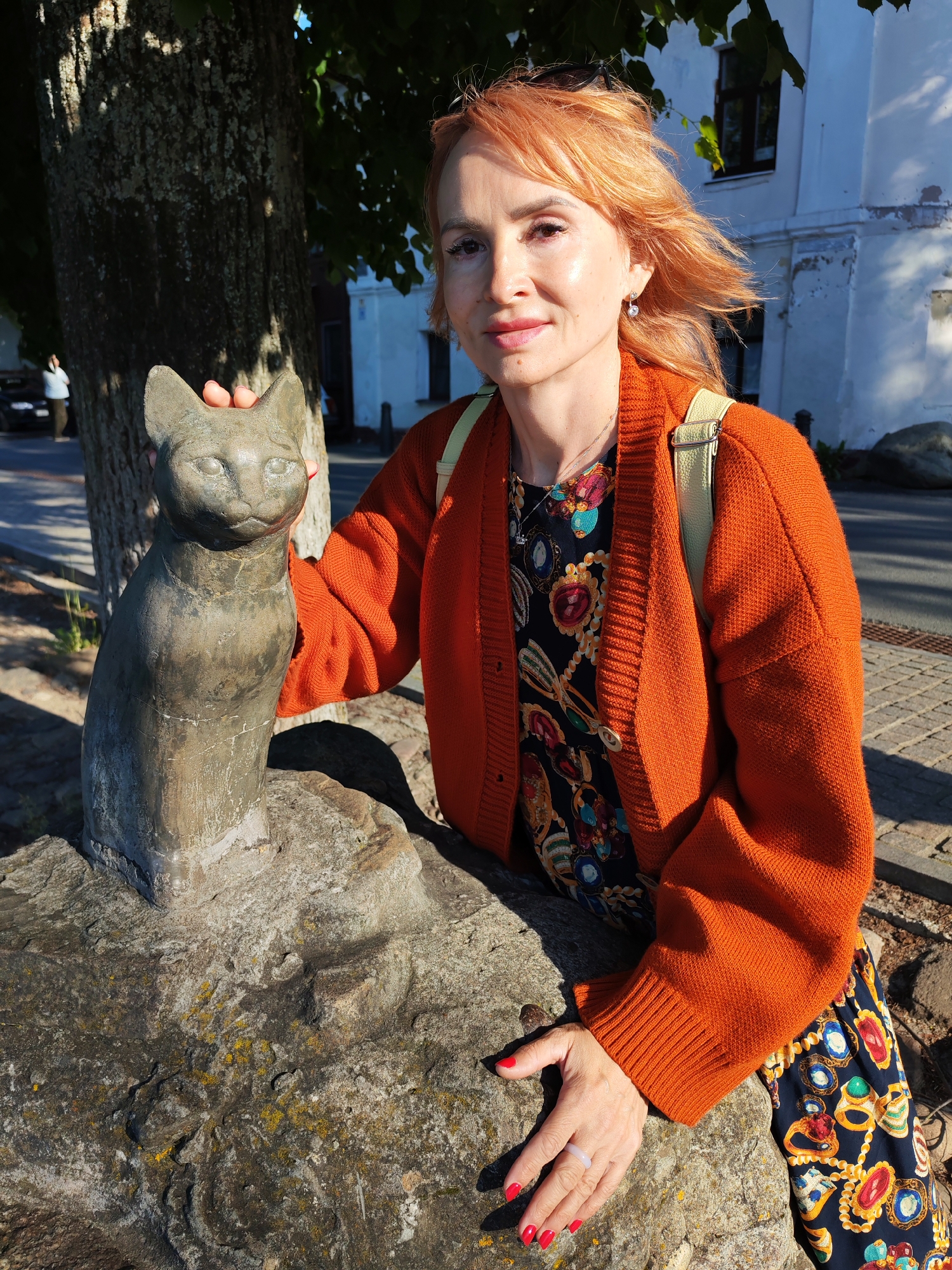 A woman with red hair poses for a photo outdoors next to a stone cat sculpture. She is wearing an orange cardigan over a dark dress with a colorful jewel pattern. A light-colored backpack is slung over her shoulder. Her right hand rests on the stone sculpture, and her left hand is visible on a rock in the foreground, adorned with a ring. The sculpture is a seated cat with a weathered, metallic finish. The scene appears to be in Plyos, Russia, with a tree to the left and a white building in the background. The lighting suggests it is daytime, likely sunny, given the shadows. The woman has a pleasant expression.