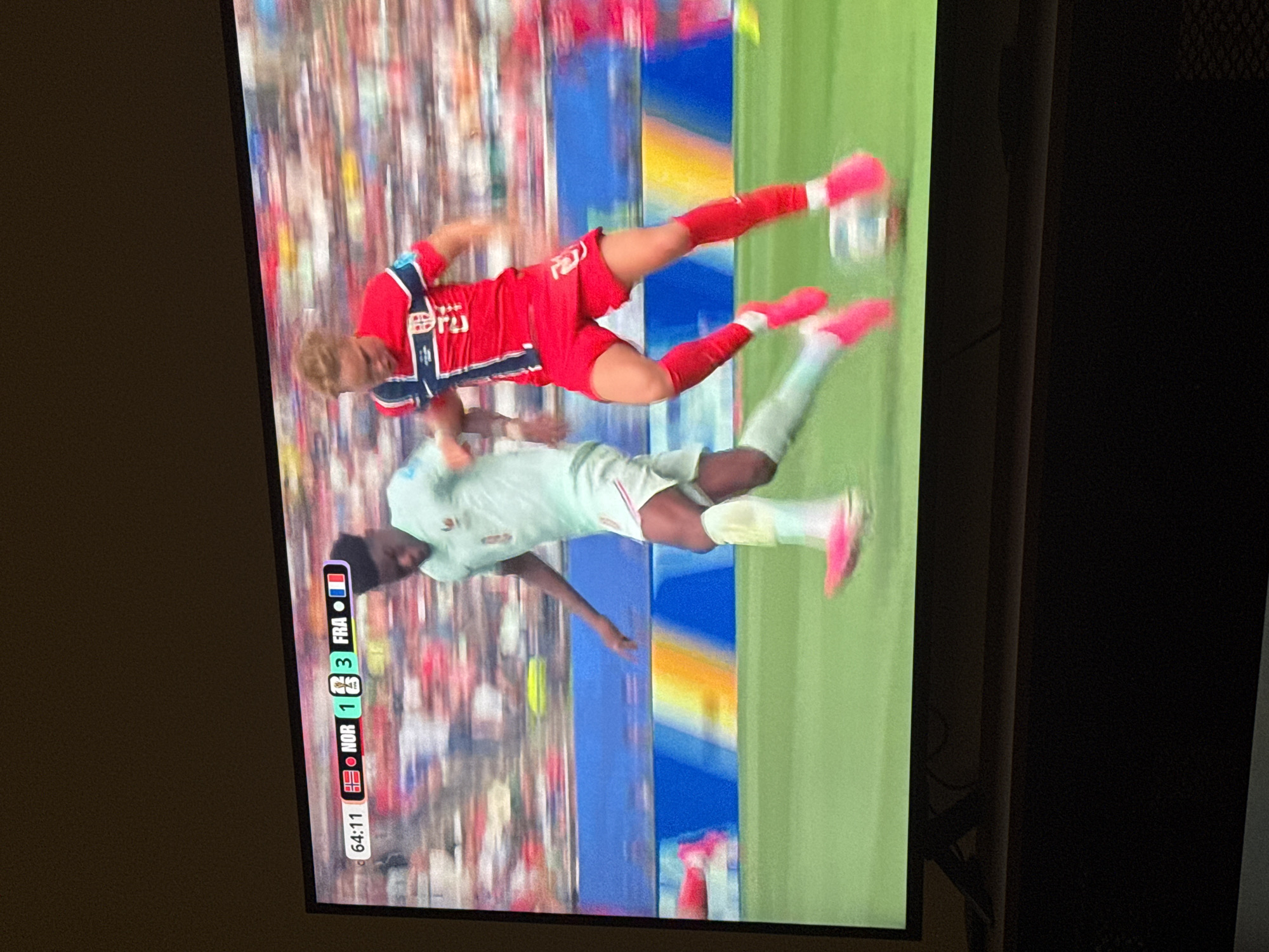 This is a screenshot from a television screen, showing a soccer match. Two male soccer players are in motion on a green field. One player, wearing a red jersey with white and blue trim and red shorts, is in mid-air, appearing to kick a soccer ball. He has blond hair and is wearing pink cleats. The other player, wearing a light blue jersey and light blue shorts, is also in motion, seemingly defending or reacting to the play. He has dark skin and hair and is wearing pink cleats. The background shows a blurred stadium filled with spectators.
Along the bottom left of the screen, a scoreboard graphic is visible. It displays "64:11" which likely indicates the current game time. To the right of the time are two national flags and scores. The first shows a white flag with a red cross, labeled "NOR" (Norway), with a score of "1". The second shows a blue, white, and red tricolor flag, labeled "FRA" (France), with a score of "3".
The image is rotated 90 degrees counter-clockwise. The lighting appears to be natural, suggesting an outdoor daytime event. The setting is a soccer stadium, and the event is a soccer match between Norway and France. The players' dynamic poses suggest an active moment in the game.