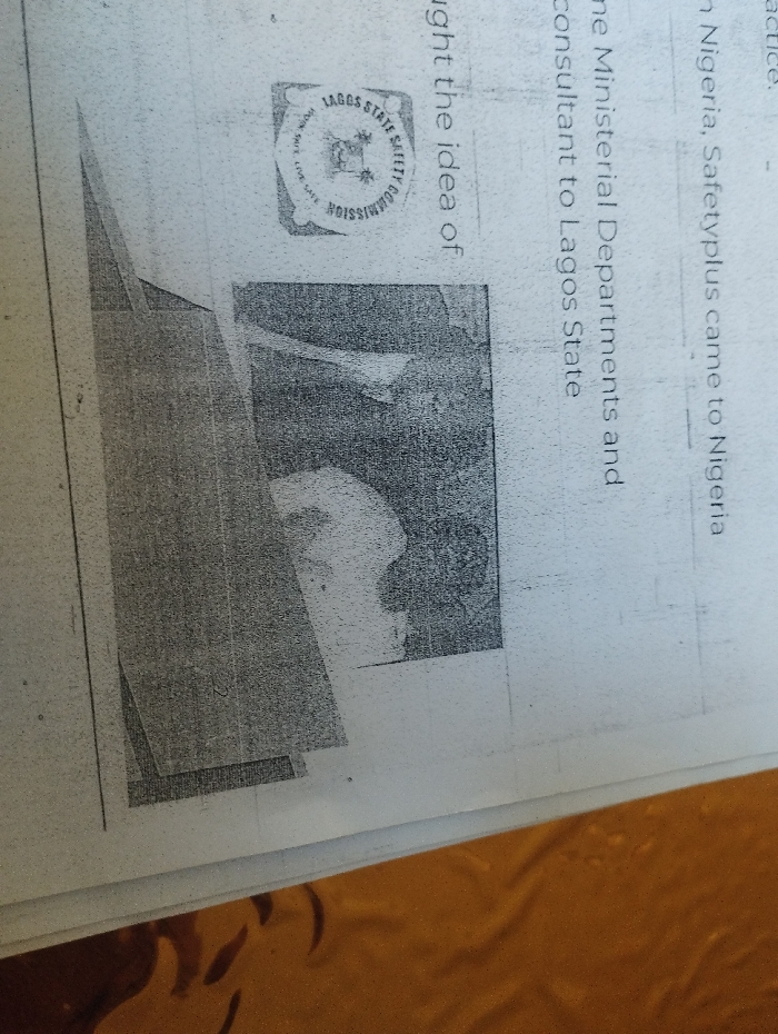 The image shows a close-up of a page from a document with a grainy, pixelated appearance. It depicts a section of text, the word "commission", and a graphic image within a rectangular box. The image is difficult to discern as it is blurred. The text visible in the image reads "the idea of the Ministerial Departments and consultant to Lagos State". The coordinates provided do not correspond to any specific location in Ashaka, Nigeria. It is likely the image is from a different document. The document is printed on white paper. The bottom of the document is partially visible, and a section of a gold-colored surface is visible beneath it. There are no identifiable individuals, subjects, objects, or actions in the image.