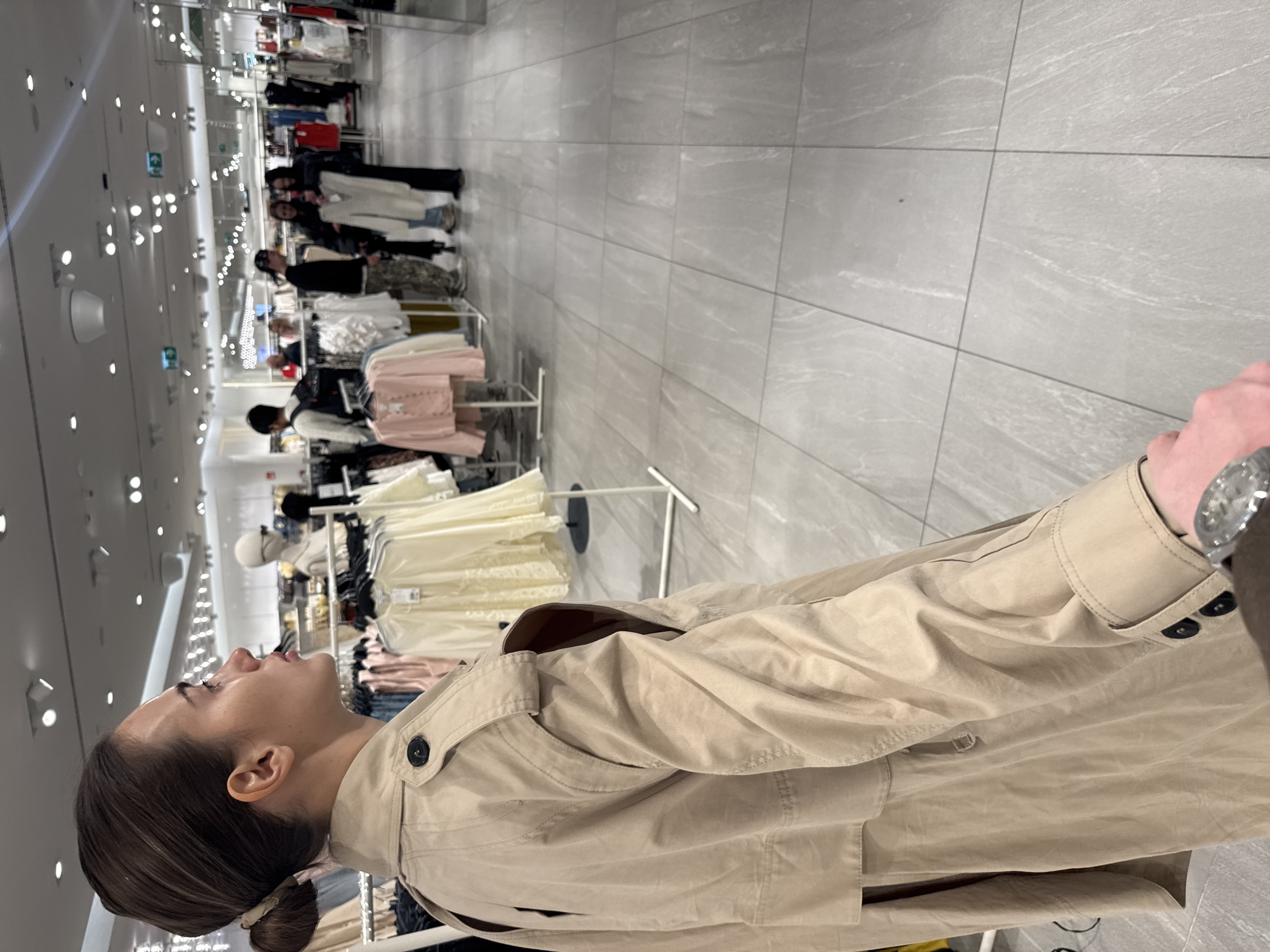 A woman wearing a tan trench coat is looking up at a rack of clothes in a store. The store has grey tile floors and bright lights. There are other people browsing clothes in the background. The city and the specific location of the store are not evident in the picture.
