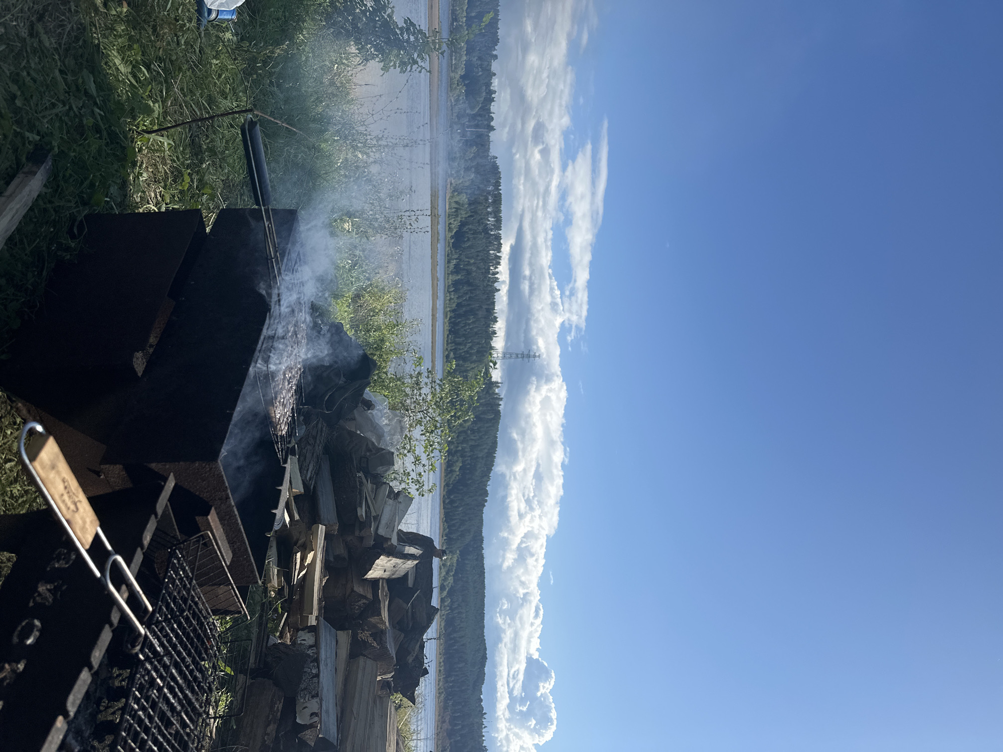An outdoor scene depicts a barbecue pit on the grassy bank of a wide river. A metal grill is filled with sizzling meat, emitting plumes of smoke that drift towards the water. In the background, a dense forest lines the opposite bank, with a power transmission tower visible in the distance. Piles of chopped firewood are stacked to the right of the grill, suggesting preparations for a long cookout. The sky above is a vibrant blue, dotted with fluffy white clouds, indicating pleasant weather. The scene appears to be located in Chaikovsky, Russia, known for its scenic riverfront and abundant natural beauty.