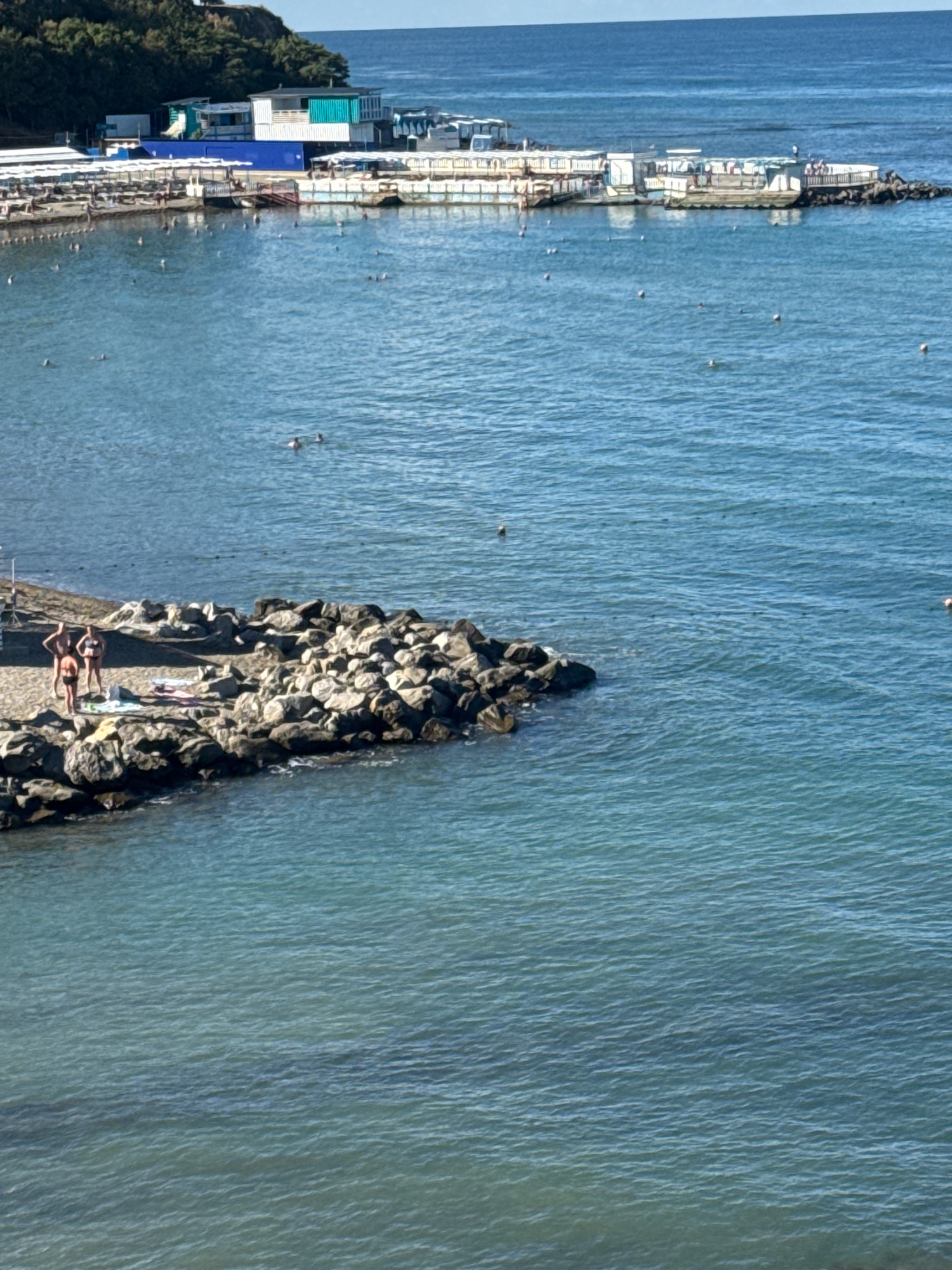 An aerial shot captures a sunny day at a beach resort in Anapa, Russia. In the foreground, a rocky jetty juts into the clear blue sea. Three people in swimwear are standing on the sandy area of the jetty, with beach towels laid out on the rocks. Further out in the water, several buoys and some individuals swimming are visible. In the background, a long, white, pier-like structure extends into the water, lined with rows of beach umbrellas and lounge chairs, indicating a developed resort area. Buildings, including a bright blue and white structure, are situated on the pier, and beyond that, a forested hillside rises against the horizon. The sea is calm and reflects the bright sky.