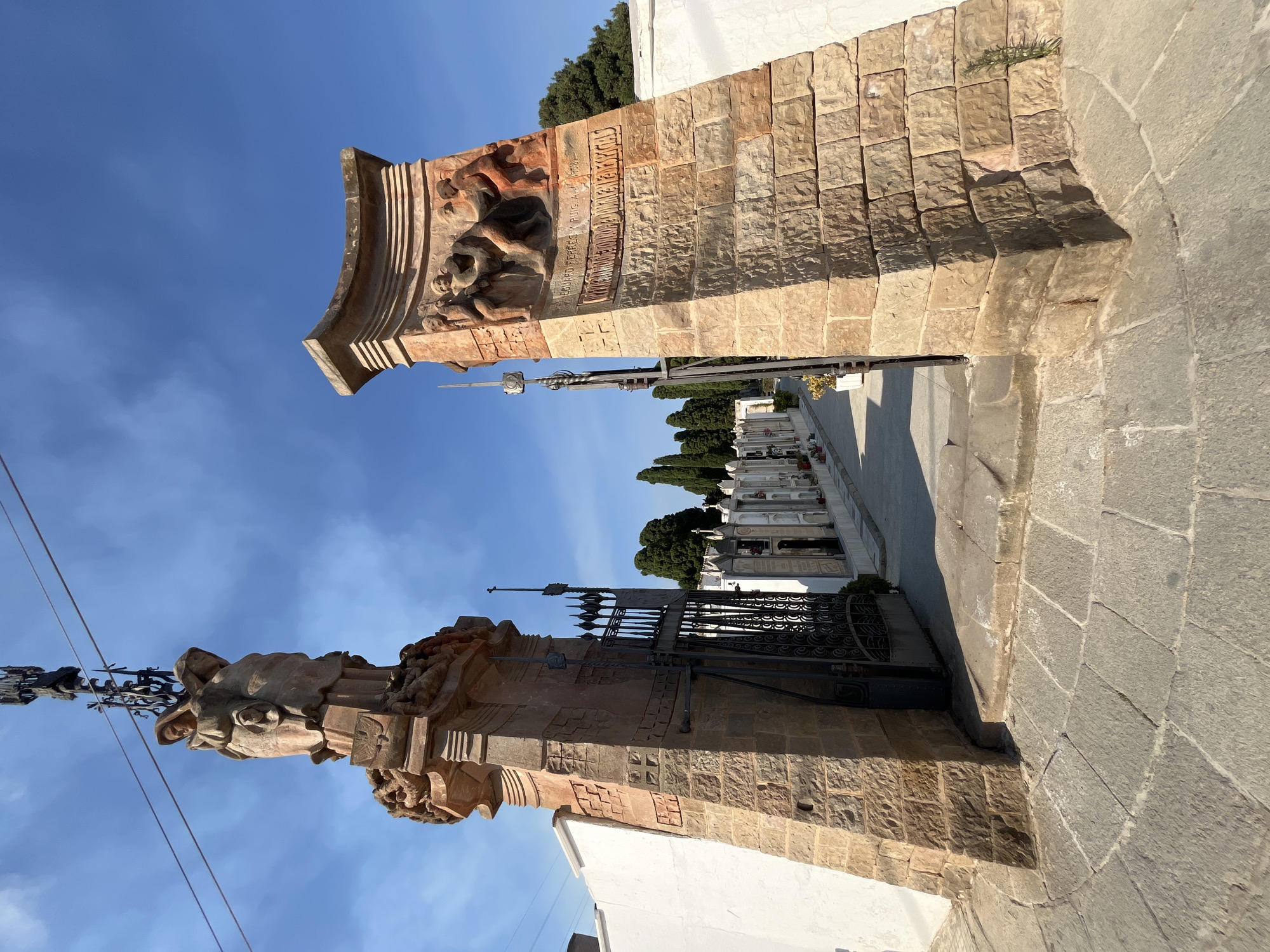 The image shows the entrance to the Vilassar de Mar cemetery in Spain. The entrance is flanked by two large stone pillars. The pillar on the left is topped with a sculpture of two figures and an ornate metal structure. The pillar on the right features a relief sculpture of figures. A black metal gate stands open between the pillars, leading into the cemetery. Beyond the gate, rows of white mausoleums and tombstones are visible, with trees lining the paths. The ground in front of the entrance is paved with cobblestones. The sky is blue with some wispy clouds.