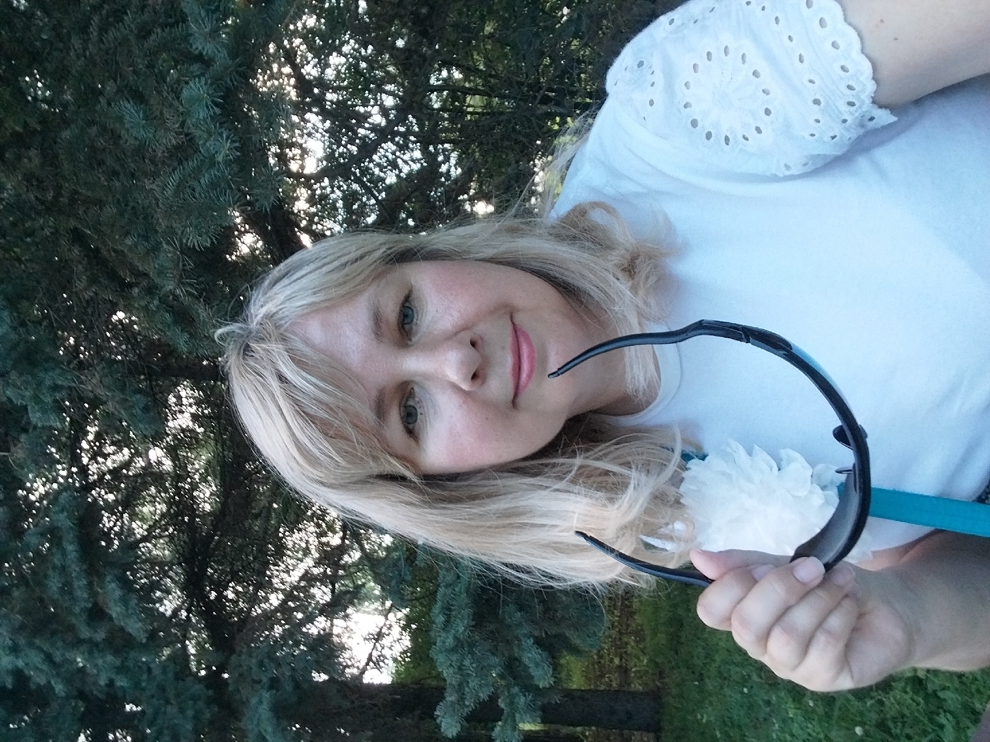A woman with blonde hair and fair skin is taking a selfie outdoors. She is wearing a white t-shirt with embroidered sleeves and holding a pair of black sunglasses. She has a white flower pinned to her shirt. The background consists of trees with green foliage and dappled sunlight. The image appears to be taken during the daytime.