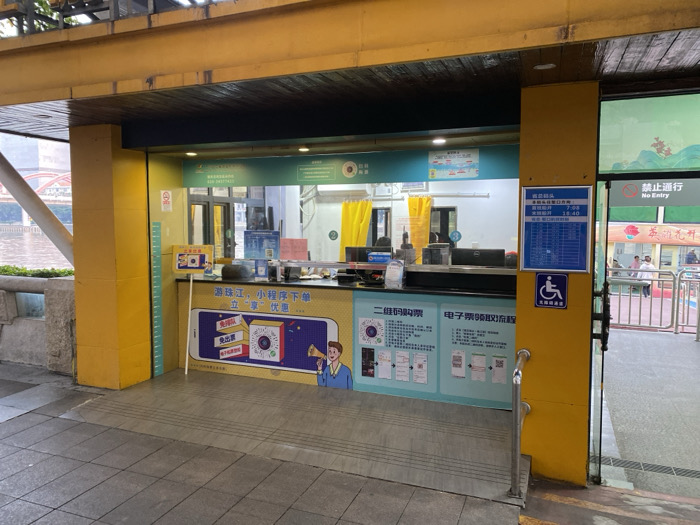 This media file captures an outdoor ticket booth, likely for a boat tour or ferry, given the visible body of water in the background. The booth features a counter with monitors and various signs, all housed under a yellow structural frame with a wooden ceiling. The ground in front appears wet, suggesting an overcast day or recent rain.
The main activity indicated is ticket purchasing and information dissemination. A large graphic on the counter advertises "游珠江, 小程序下单 立享优惠..." (Pearl River Tour, order via mini-program to enjoy discounts...). It also highlights "免出票" (Paperless/No ticket printing) options. Sections for "二维码购票" (QR code ticket purchase) and "电子票领取流程" (E-ticket collection process) are prominently displayed with QR codes and instructions.
A blue sign to the right of the counter lists departure times, with "08:00" and "18:40" clearly visible. Below this, a wheelchair accessibility symbol is affixed to a yellow column. Beyond the booth, metal railings lead to an area marked with a "禁止通行" (No Entry) sign, where a few individuals are visible in the background. The scene suggests a public transport or tourist facility in Guangzhou, China.