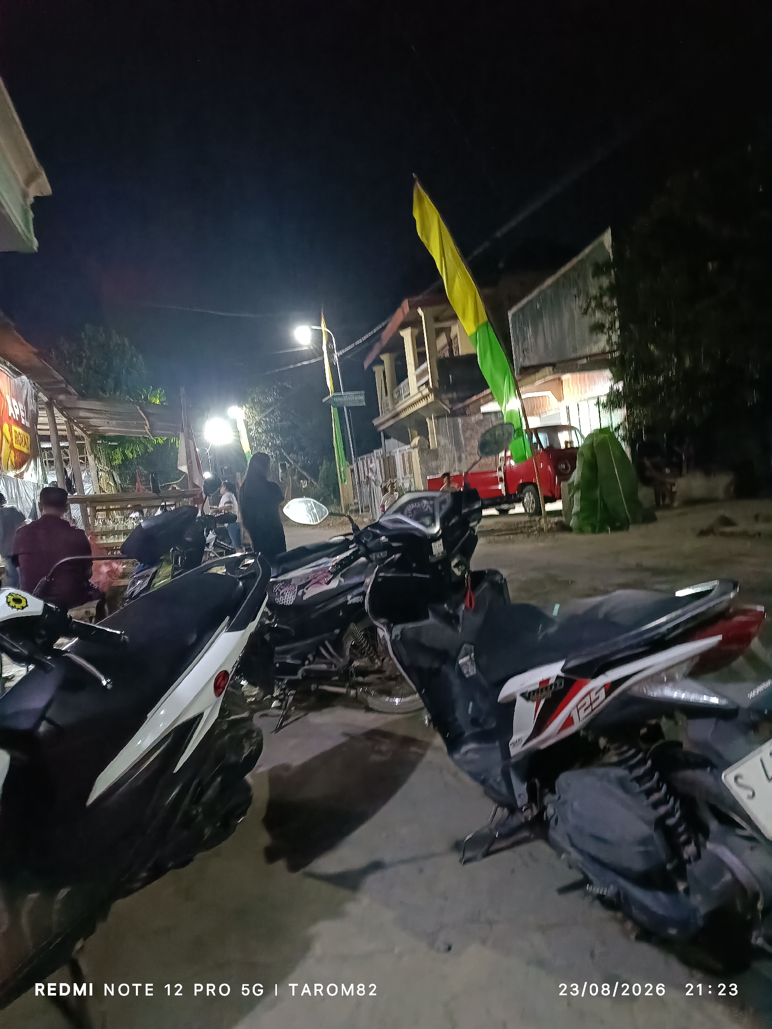 The image depicts an outdoor scene at night in what appears to be a marketplace or a street in Rejoagung, Indonesia. Several motorcycles are parked, with two in the foreground being the most prominent. The motorcycle on the left is white and black, and the one on the right is black with red and white accents, featuring the number "125" on its side. In the background, a red truck is visible, along with a green flag and a yellow and green flag. Several individuals are present, some standing and some sitting, and structures that appear to be stalls or buildings line the street. The lighting suggests artificial illumination from streetlights and possibly from the buildings. The text overlay at the bottom of the image indicates it was taken with a "REDMI NOTE 12 PRO 5G | TAROM82" on "23/08/2026 21:23".