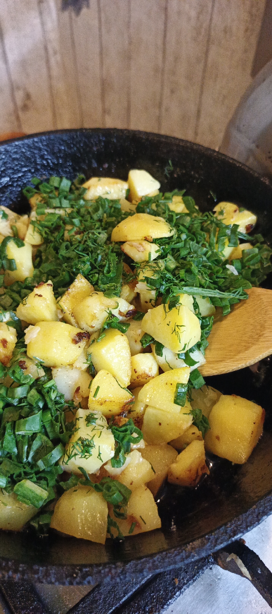 A close-up, overhead shot shows a rustic cast-iron skillet filled with golden-brown, diced potatoes that have been pan-fried. Freshly chopped green herbs, likely dill and chives, are generously sprinkled over the potatoes. A wooden spoon is partially visible on the right side of the frame, suggesting the dish is being served or stirred. The background consists of a textured, wooden wall with vertical planks, indicative of a home kitchen or a rustic restaurant setting. There are no people visible in the image. The scene captures a simple, home-style meal, common in many Russian households, including those in Chaikovsky.