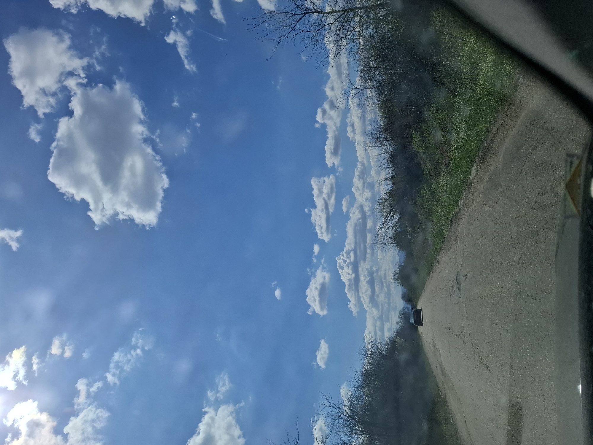 A view from inside a car traveling on a rural road. The sky is blue with scattered white clouds. Vegetation lines both sides of the road, with trees and some green growth visible. A car is traveling in the same direction ahead on the road. The exact location in Gramada, Bulgaria cannot be determined without further information.