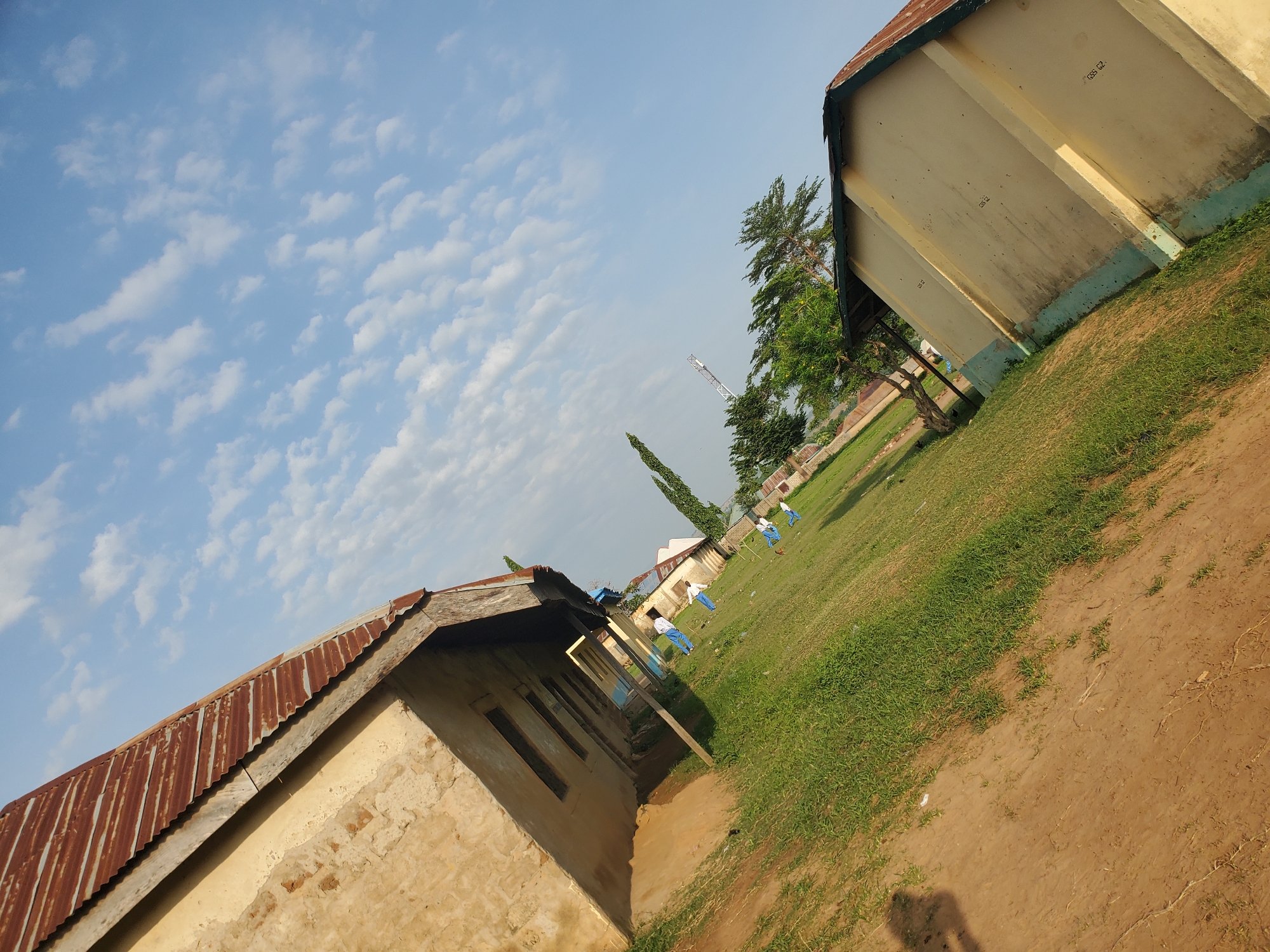 The image displays an exterior view of a school building under a clear blue sky with scattered white clouds. The building, which appears aged, has a corrugated metal roof and light-colored walls. The foreground is a mix of dirt and grass, with some shadows indicating the presence of objects or structures not fully in frame. Several individuals, likely students in uniform, are visible on the grassy area in front of the building, some walking or standing. In the background, trees and more buildings can be seen, suggesting a rural or suburban setting. The perspective is tilted, giving a dynamic feel to the composition. The scene appears to be set in Winters, United States of America.