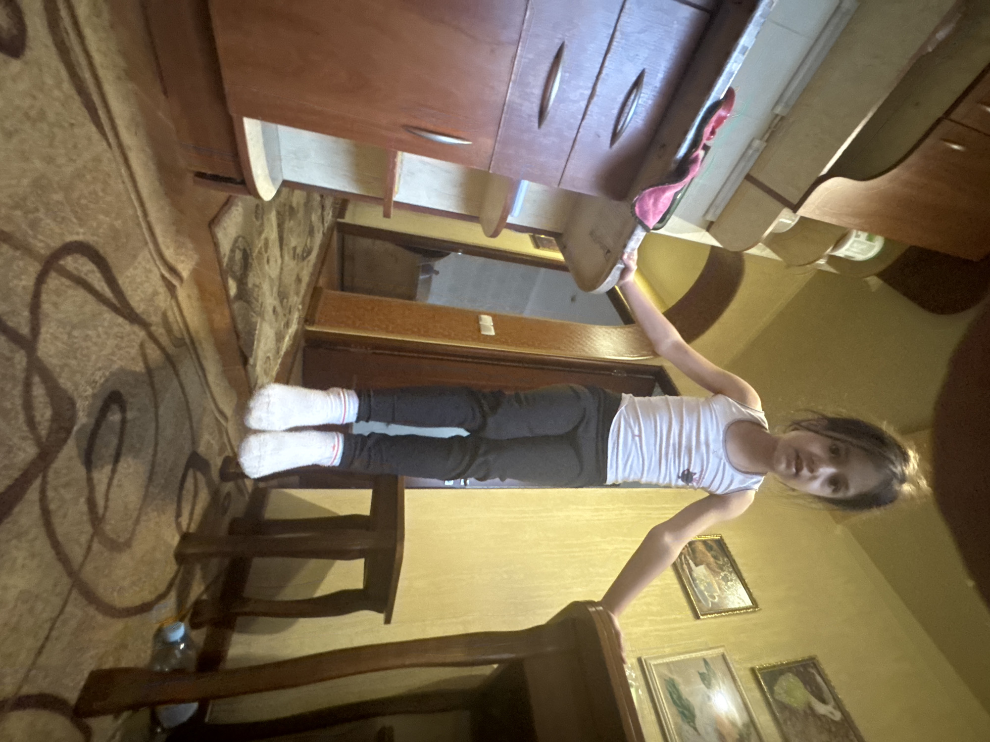 A young girl is captured indoors, appearing to stand on a wooden stool or chair, her head and upper body positioned higher than the surrounding furniture. She has dark hair and is wearing a white ribbed tank top and dark pants with white socks that have thin red and blue stripes near the cuffs. Her arms are raised, and she is holding up a light-colored, possibly wooden, rectangular object, which has a pink cloth or oven mitt draped over one end. Her gaze is directed towards the camera, showing a neutral to slightly smiling expression.
The setting is an interior space, likely a home, featuring warm, yellowish-colored walls and dark brown wooden furniture. Above the girl's head are wooden cabinets or wall-mounted shelves with visible drawers and handles. Below these cabinets, there is a reflective surface, possibly a countertop or a window. A patterned rug with dark, swirling designs covers the floor to the left, where a clear plastic water bottle with a blue cap is also visible near the base of the wooden stool. Several framed pictures or paintings with dark frames are hung on the wall to the right. The lighting suggests an indoor scene, either during the day or evening. There is no discernible text visible on any objects or surfaces.