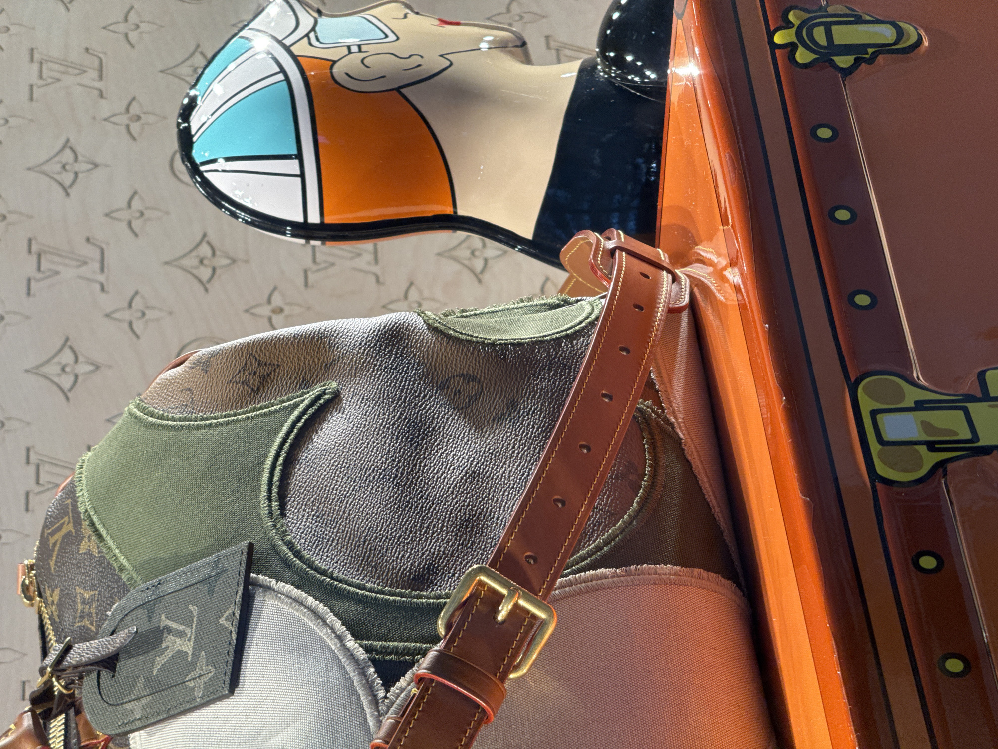 The image is a close-up, eye-level shot of a Louis Vuitton handbag with a patchwork design, featuring various textures and colors including the iconic monogram canvas, green fabric, and light grey fabric. A brown leather strap with a brass buckle is visible across the front of the bag. To the right of the bag is a mannequin head wearing a helmet with blue and white stripes and an orange and black racing suit, with a detailed profile of the face visible. Both the bag and the mannequin are placed on an orange, glossy surface, possibly a display case or the lid of a trunk, which has black and yellow decorative elements resembling hardware and clasps. The background is a light-colored wall embossed with the Louis Vuitton monogram pattern. There are no people present in the image, and no actions are being depicted. The scene appears to be set in a retail environment, likely a Louis Vuitton boutique, and based on the provided location, it is situated within Nong Prue Subdistrict, Thailand.