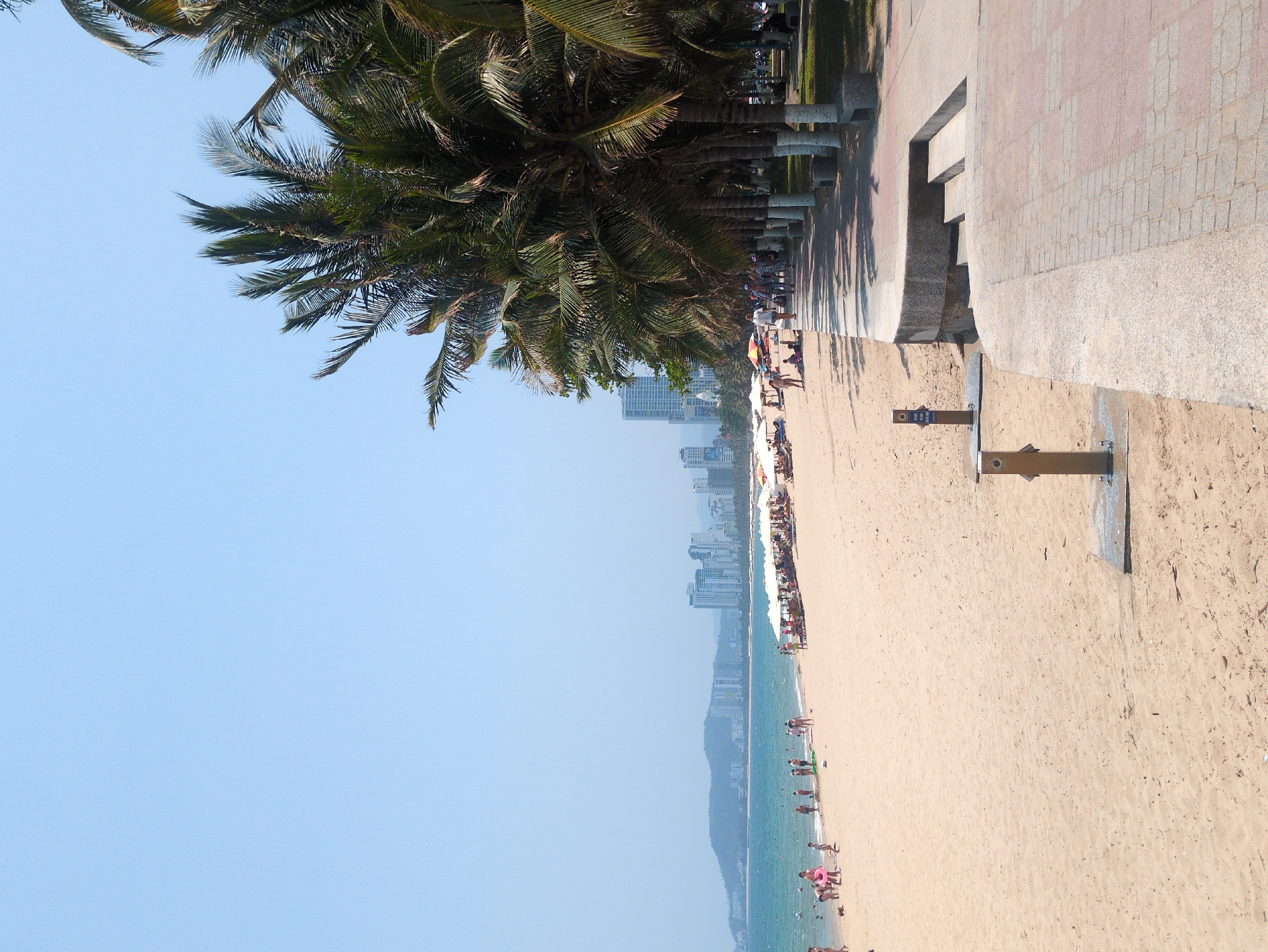 The image depicts a beach in Nha Trang, Vietnam. The scene shows a sandy beach with people sunbathing, swimming, and strolling along the shore. The clear blue sky contrasts the turquoise sea. A row of palm trees lines the edge of the beach, next to a tiled walkway with benches. A few beach showers are visible near the walkway. Buildings rise in the background, indicating an urban area.