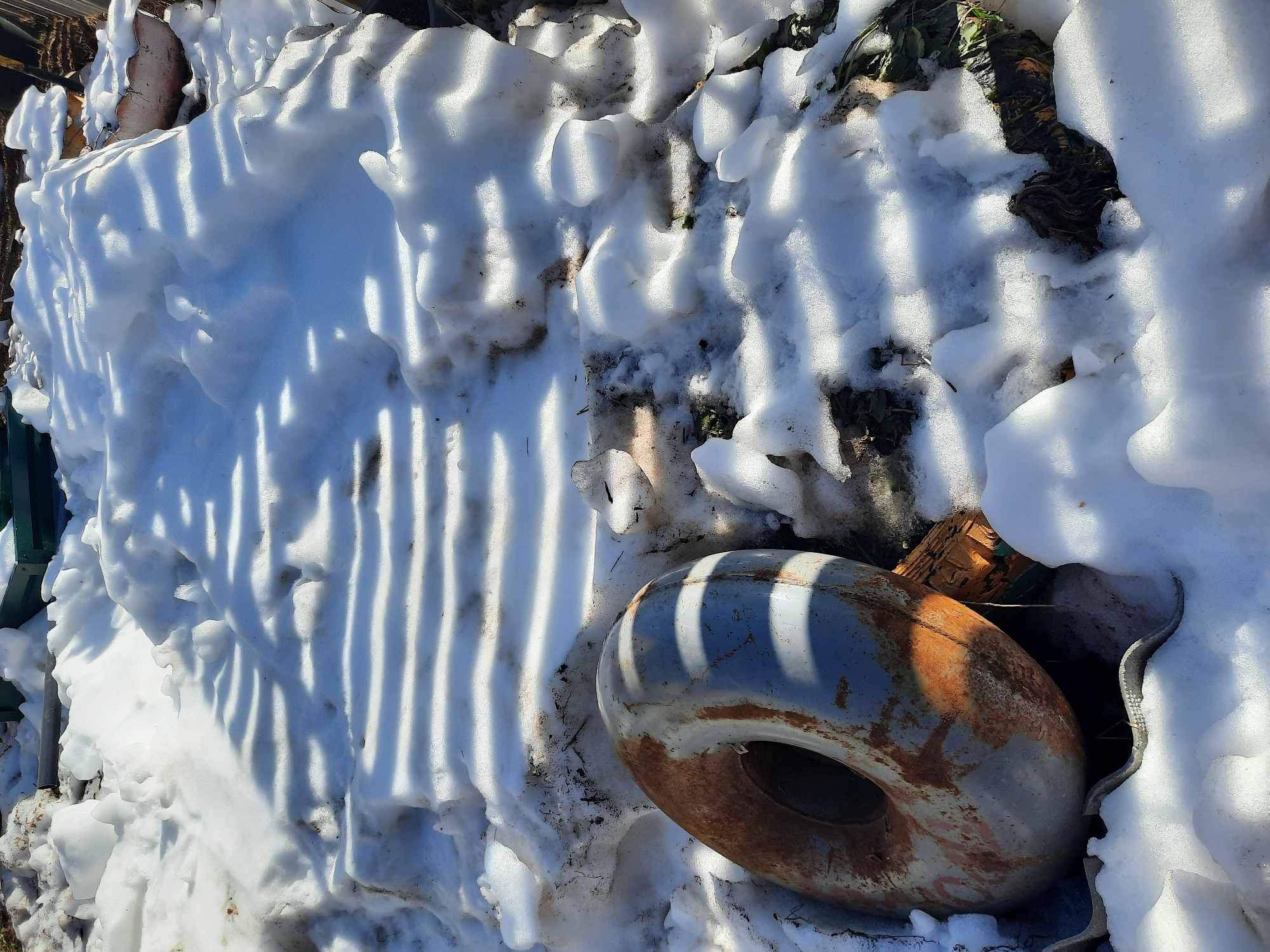 The image depicts a snowy scene, likely outdoors, in or near Krasnogorskoye, Russia, though specific identifiers beyond the general winter environment are absent. A large, donut-shaped object rests on the snow. The object is metal, primarily painted light grey, and exhibits significant rust. The snow cover is uneven, with variable depth and some exposed ground. The shadows cast across the scene suggest a structure with vertical bars, possibly a fence or railing. The scene includes other debris, visible under the snow.