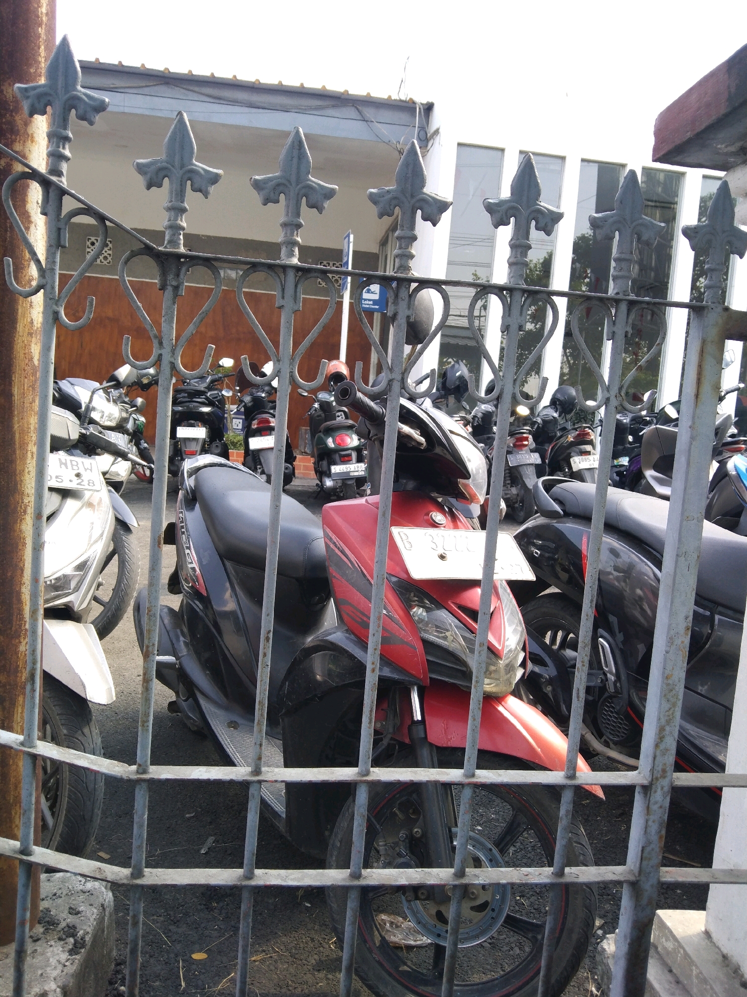 The image shows a close-up view of a weathered, grey metal fence with decorative finials and heart-shaped scrolling. Through the fence, a row of parked motorcycles is visible, with the foreground dominated by a red and black scooter. The scooter has a license plate with the text "B 3272 L", a white tag below it, and a red front fender. Behind it, other motorcycles are parked, some with visible license plates such as "HBW 05.28" and others with obscured details. The motorcycles are parked on an asphalt surface, and in the background, a white building with large windows and a white roof is visible. The scene suggests an outdoor parking area, possibly in Bojonegoro, Indonesia, given the style of the motorcycles and the plate format. The lighting indicates daytime.