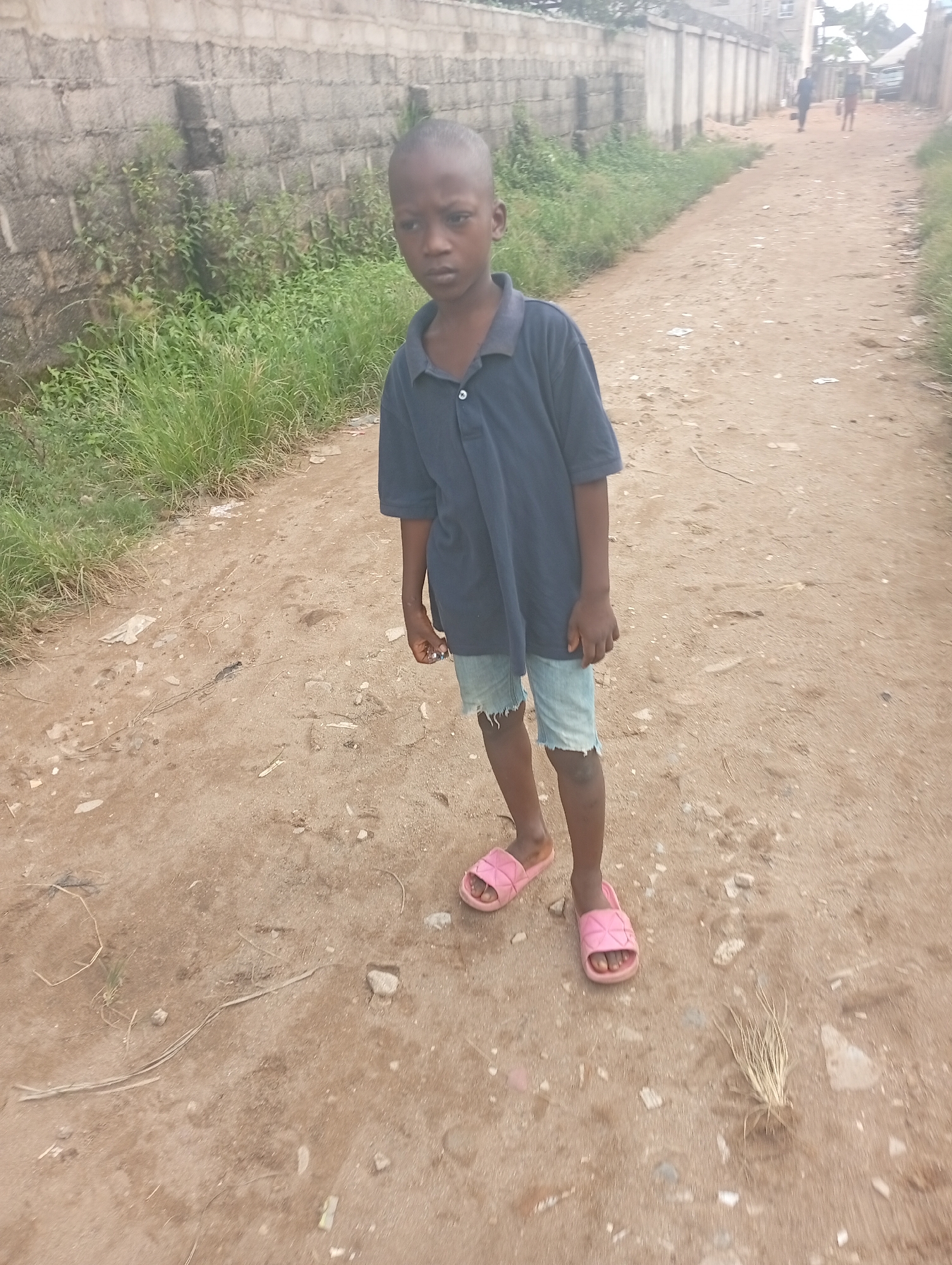 A young boy stands on a dirt road in Eziudo, Nigeria. He is wearing a navy blue polo shirt, denim shorts, and pink sandals. A concrete block wall with greenery runs along the left side of the road. Two individuals are walking in the distance on the road.