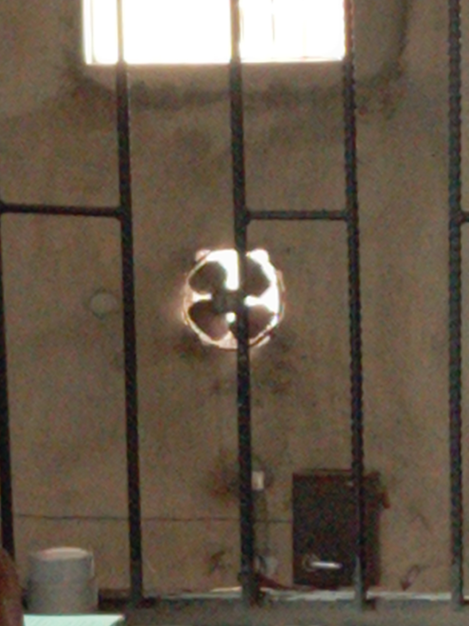 This image captures an interior view, likely of a secure or utilitarian room in Owerri, Nigeria, dominated by the presence of sturdy metal bars in the foreground.
At the top, a rectangular window emits a strong, bright light, indicating daytime. Below this window, the textured, unpainted concrete wall fills most of the background. Roughly in the center of the wall, there is a circular opening containing what appears to be an extractor fan or vent with visible blades, also illuminated from behind.
In the lower right section of the image, mounted on the wall, is a dark, rectangular box, possibly an electrical panel or a small utility box, featuring a distinct handle or latch on its front. To the lower left, a light-colored, cylindrical object rests on the floor or a low ledge. The bottom of the frame shows a horizontal bar or edge, likely part of the structure the metal bars are set into.
The scene conveys a stark and functional environment, with no people visible and no specific activity occurring. The rough concrete walls and the robust metal grille contribute to a sense of security or containment. No discernible text is visible in the image.